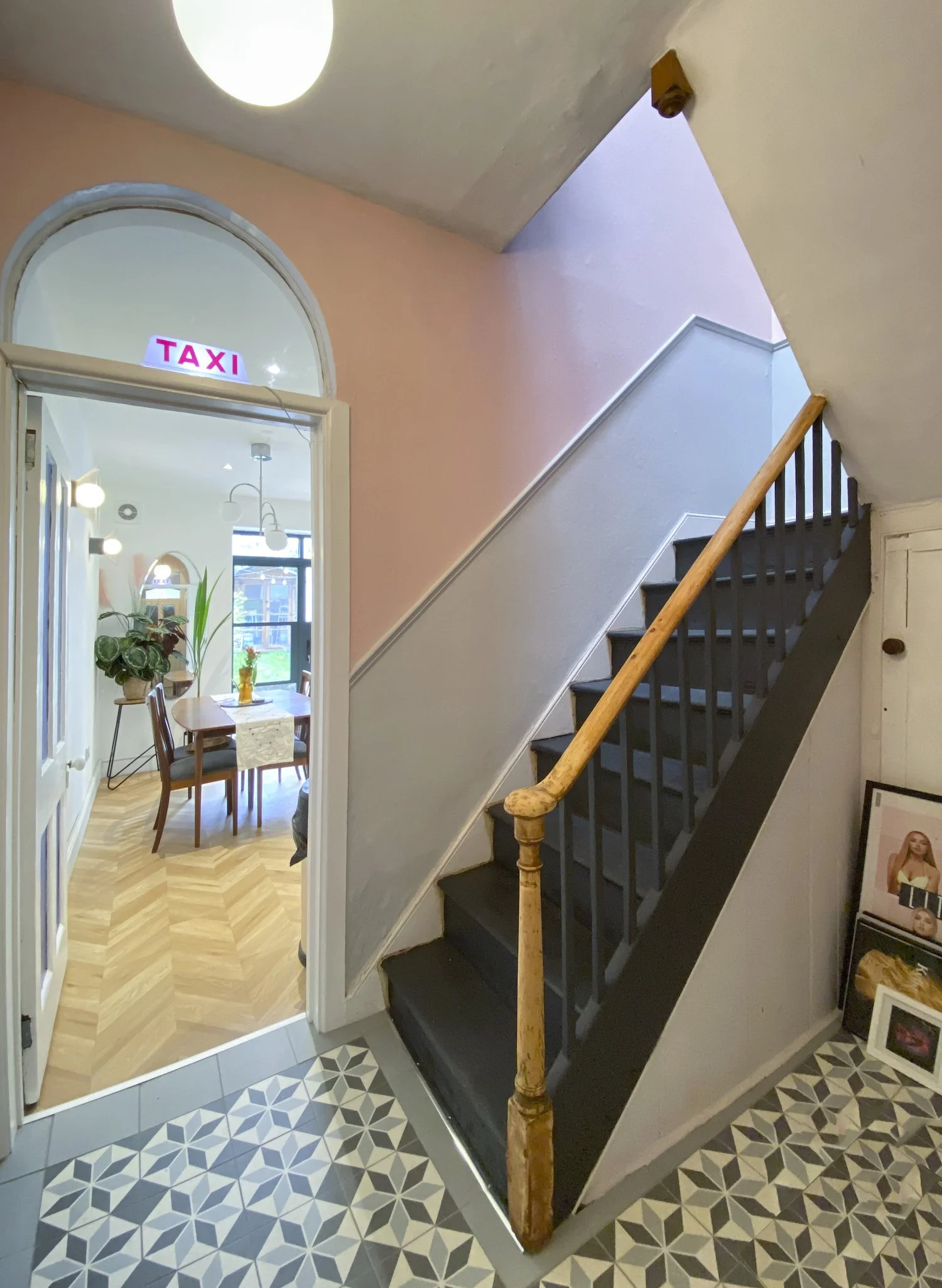 Interior view of a house showing a staircase with black steps and a wooden handrail, a patterned floor, and a doorway leading to a dining room with a pink wall and a 'TAXI' sign above the doorframe.