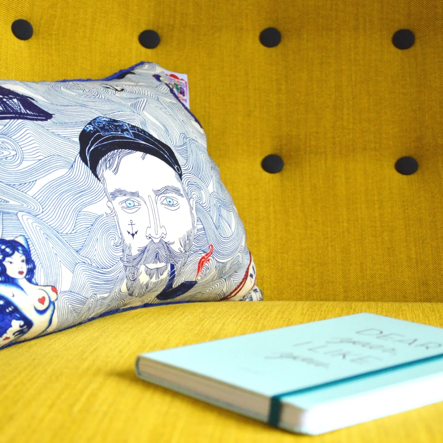 A decorative pillow with a line drawing of a bearded man wearing a beanie and a woman with dark hair and red lipstick, placed on a mustard yellow upholstered chair with black buttons. A small light blue notebook rests on the seat in front of the pillow.