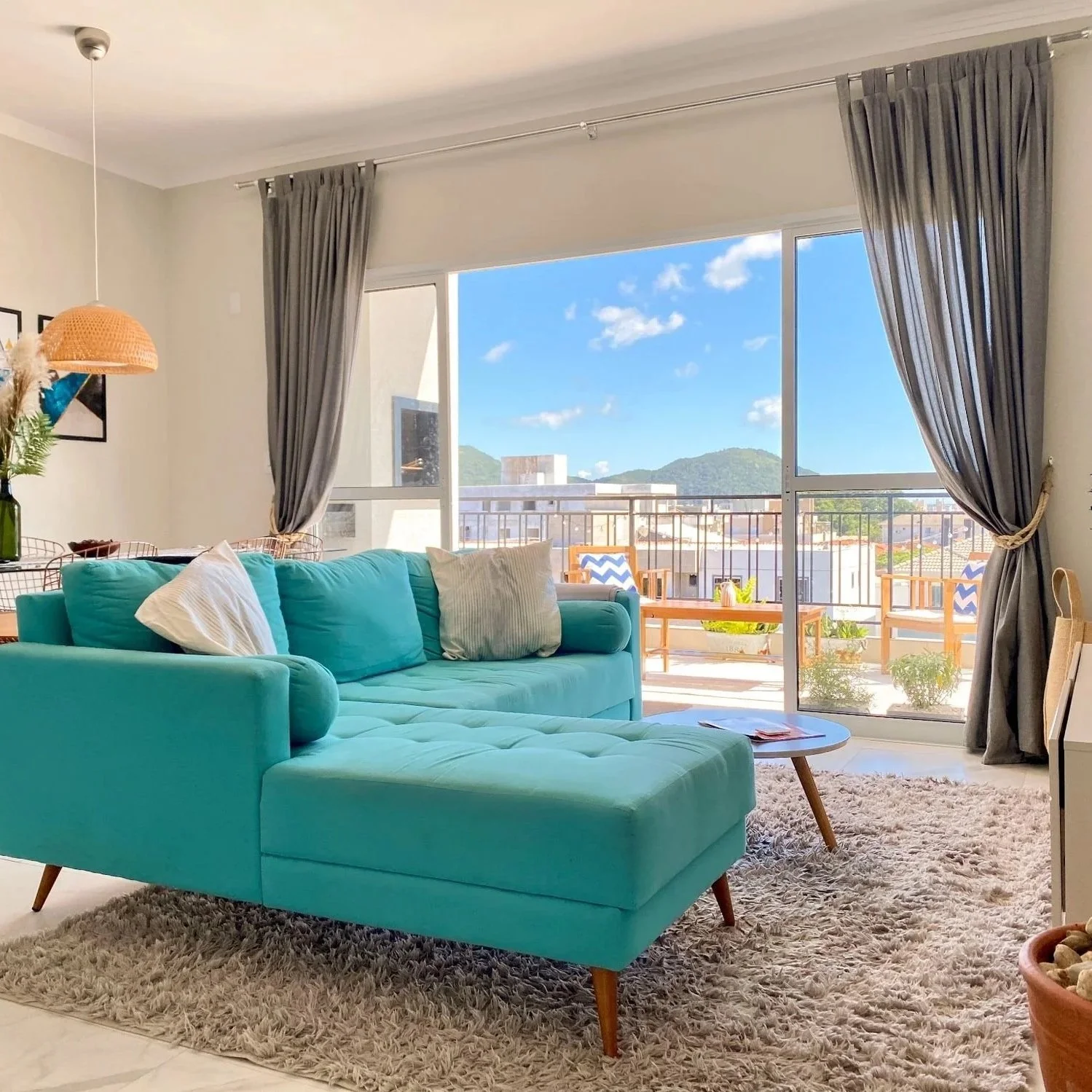 Bright living room with turquoise sectional sofa, cream and striped pillows, beige shag rug, wooden coffee table, large sliding glass door with gray curtains, balcony with chairs and potted plants, scenic mountain view outside.