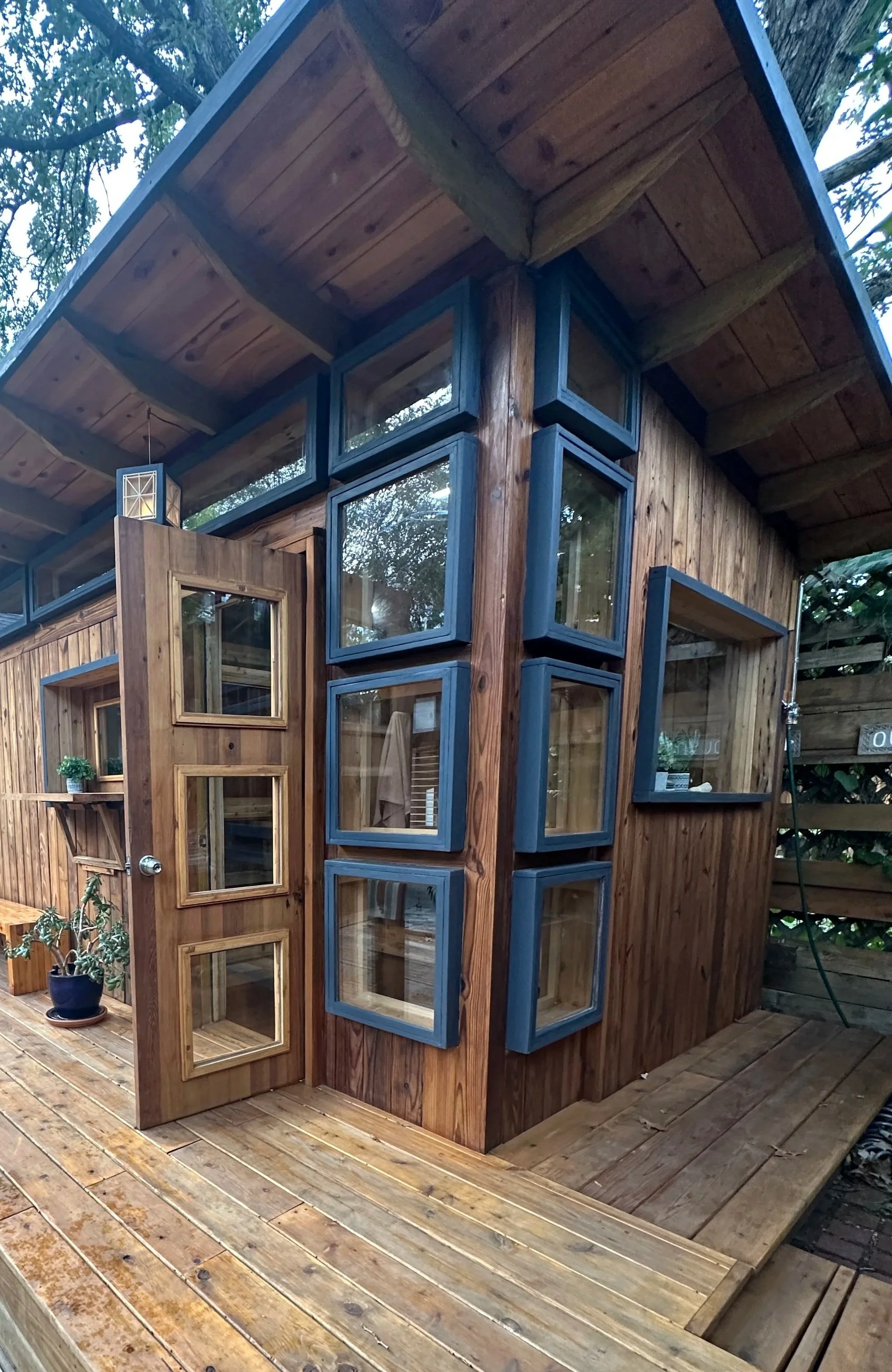 A small wooden cabin with a sloped roof, featuring multiple square and rectangular glass windows framed in dark gray, and a wooden door with glass panels. There are potted plants and a wooden deck surrounding the cabin.
