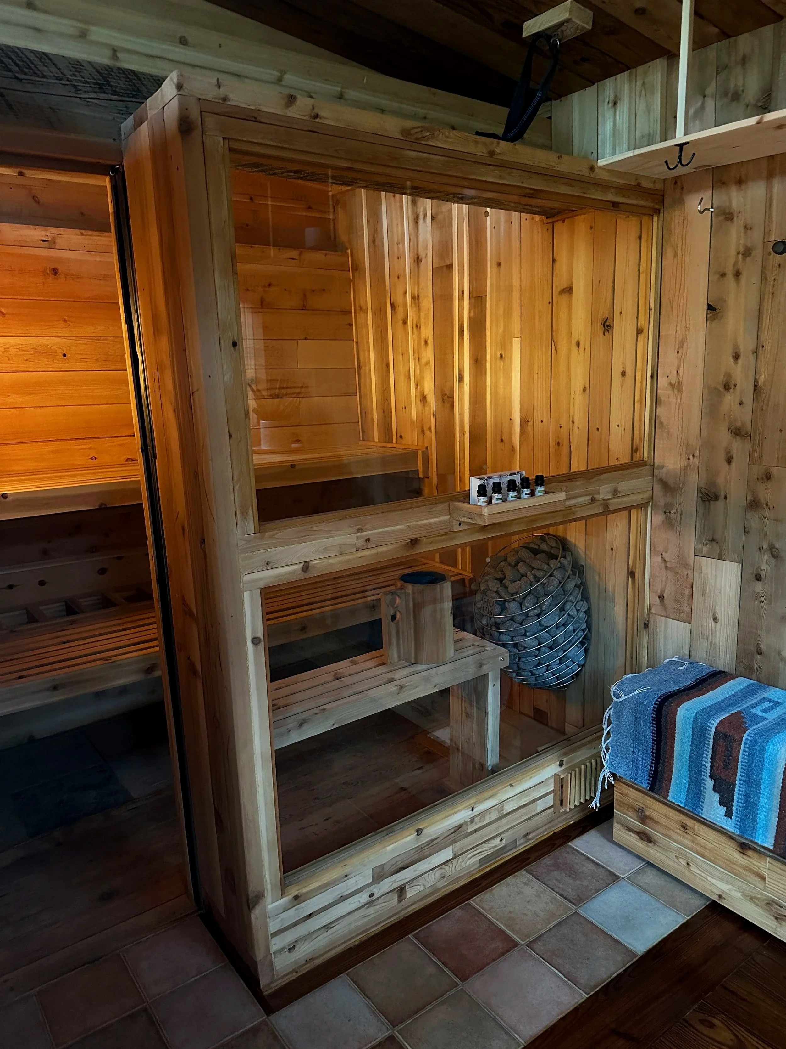 Indoor wooden sauna with glass door, a sauna bucket, a towel, and essential oils on a shelf.