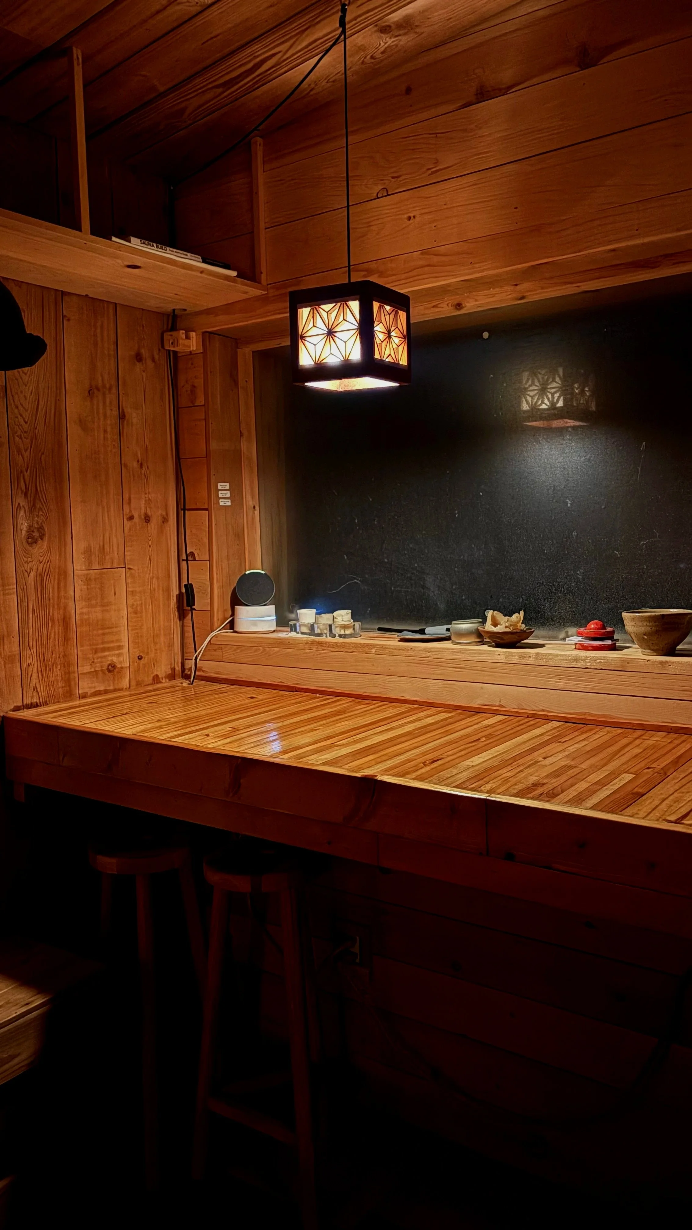 A cozy wooden kitchen space with a wooden countertop, black chalkboard wall, hanging lantern-style light, small speaker, and various dishes and utensils on the countertop.