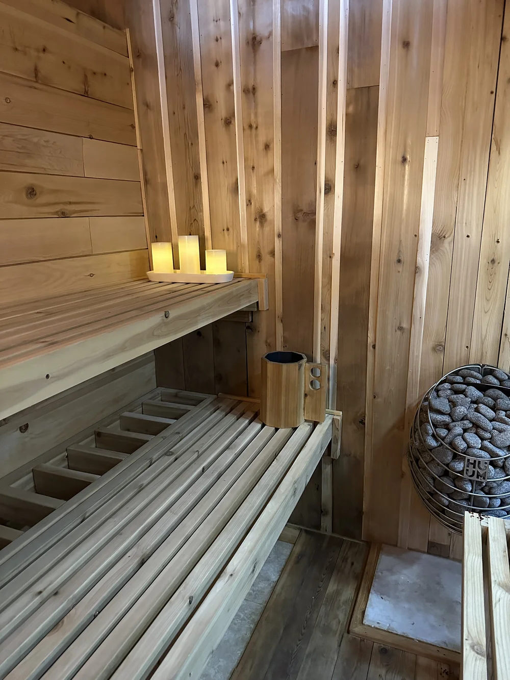 A wooden sauna interior with a lit candle arrangement on a shelf, a wooden bucket, and a sauna heater filled with stones.