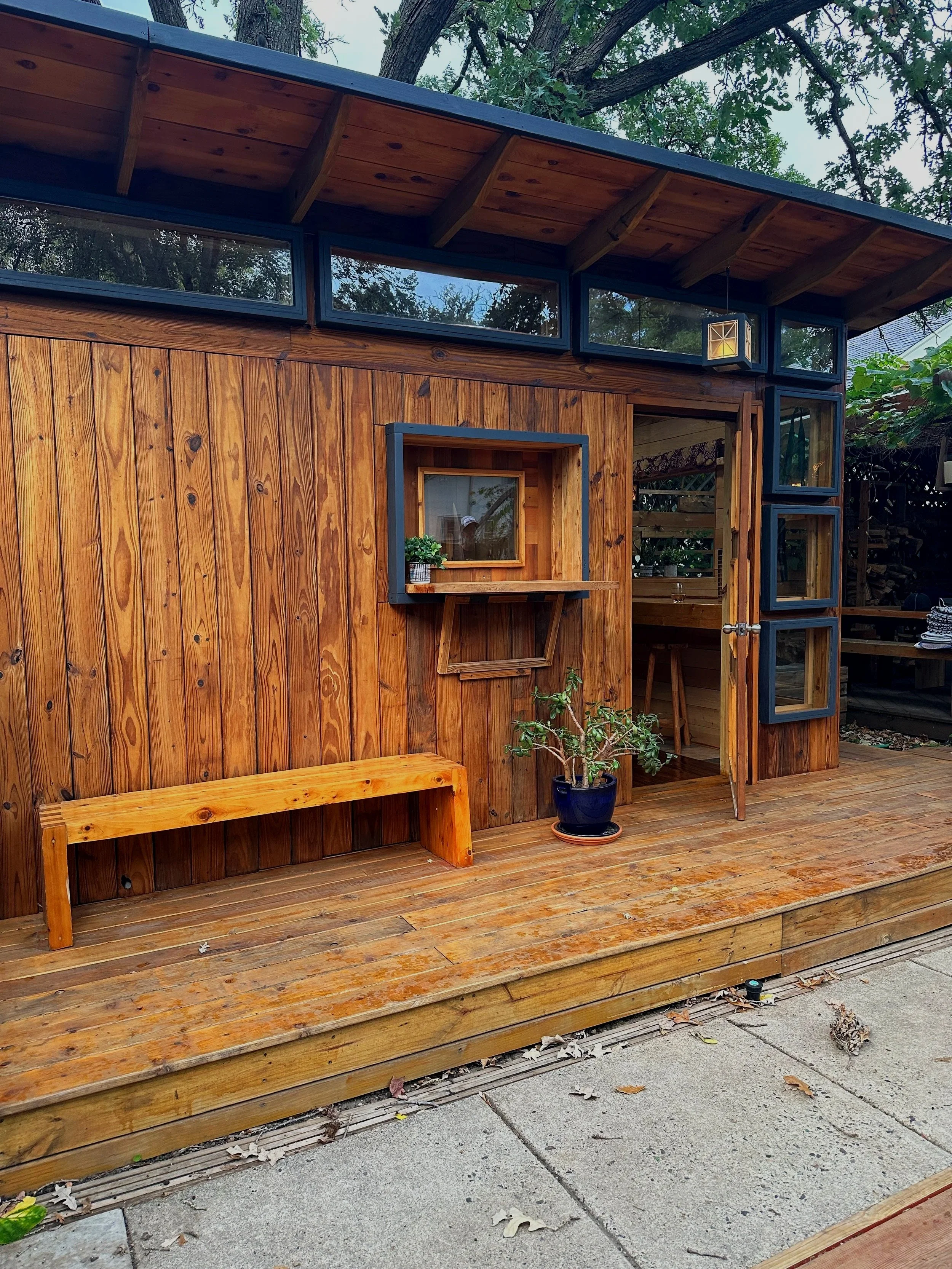 A small wooden cabin or shed with an exterior deck, featuring a bench and a potted plant outside, along with large windows and a door leading inside.