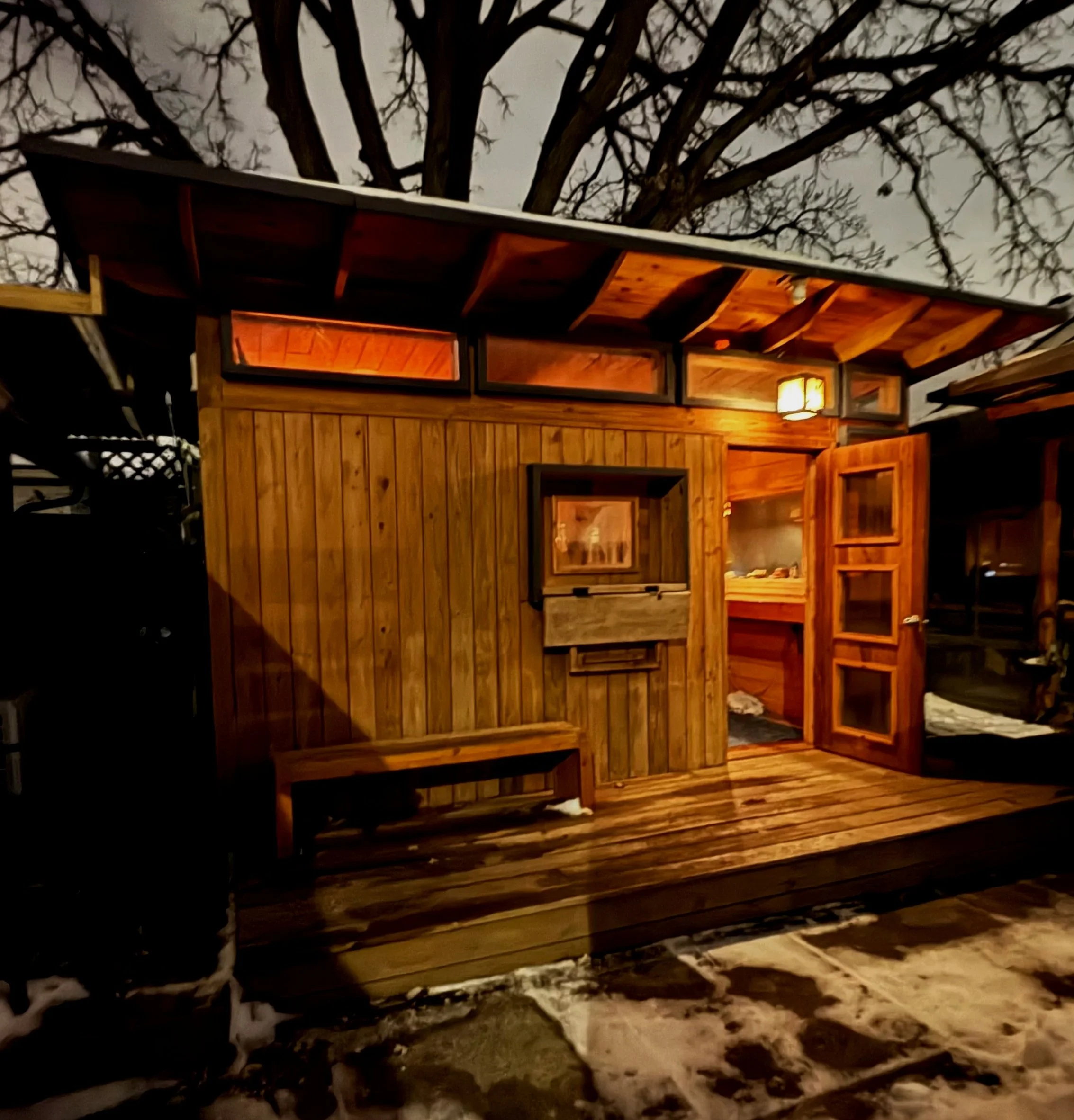A small wooden cabin with a bench outside, illuminated by a warm light, surrounded by snow at night, with tree branches overhead.