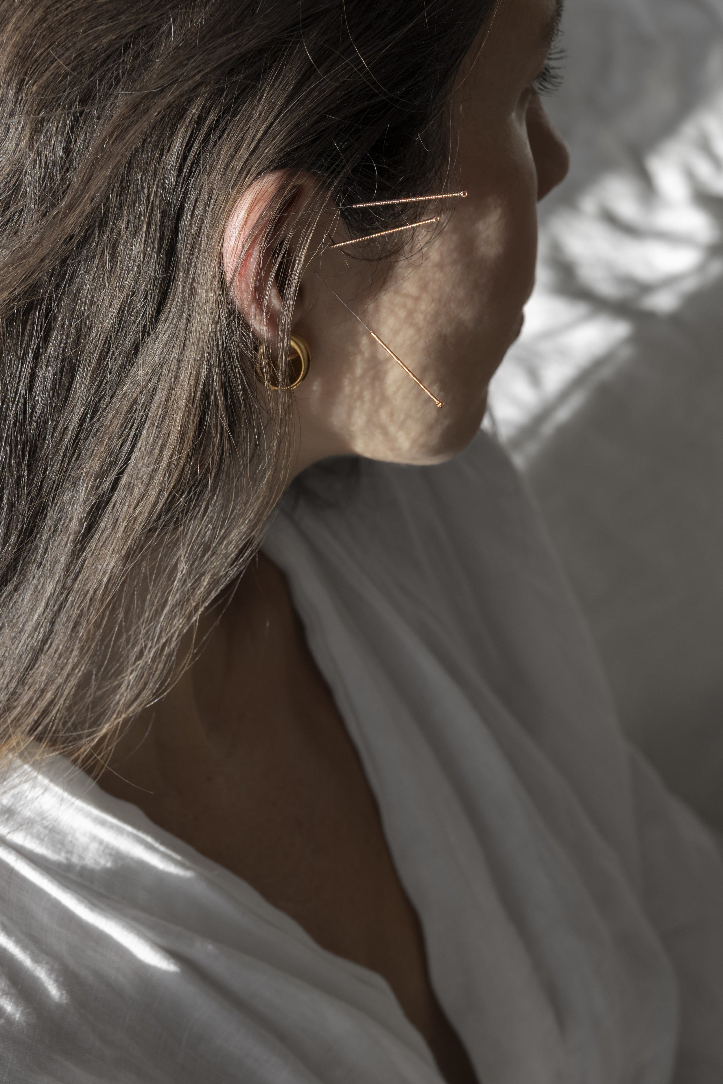 Close-up of a woman with brown hair, wearing gold earrings, with multiple pins in her ear, and a white top, sitting on a bed or couch with sunlight casting shadows.