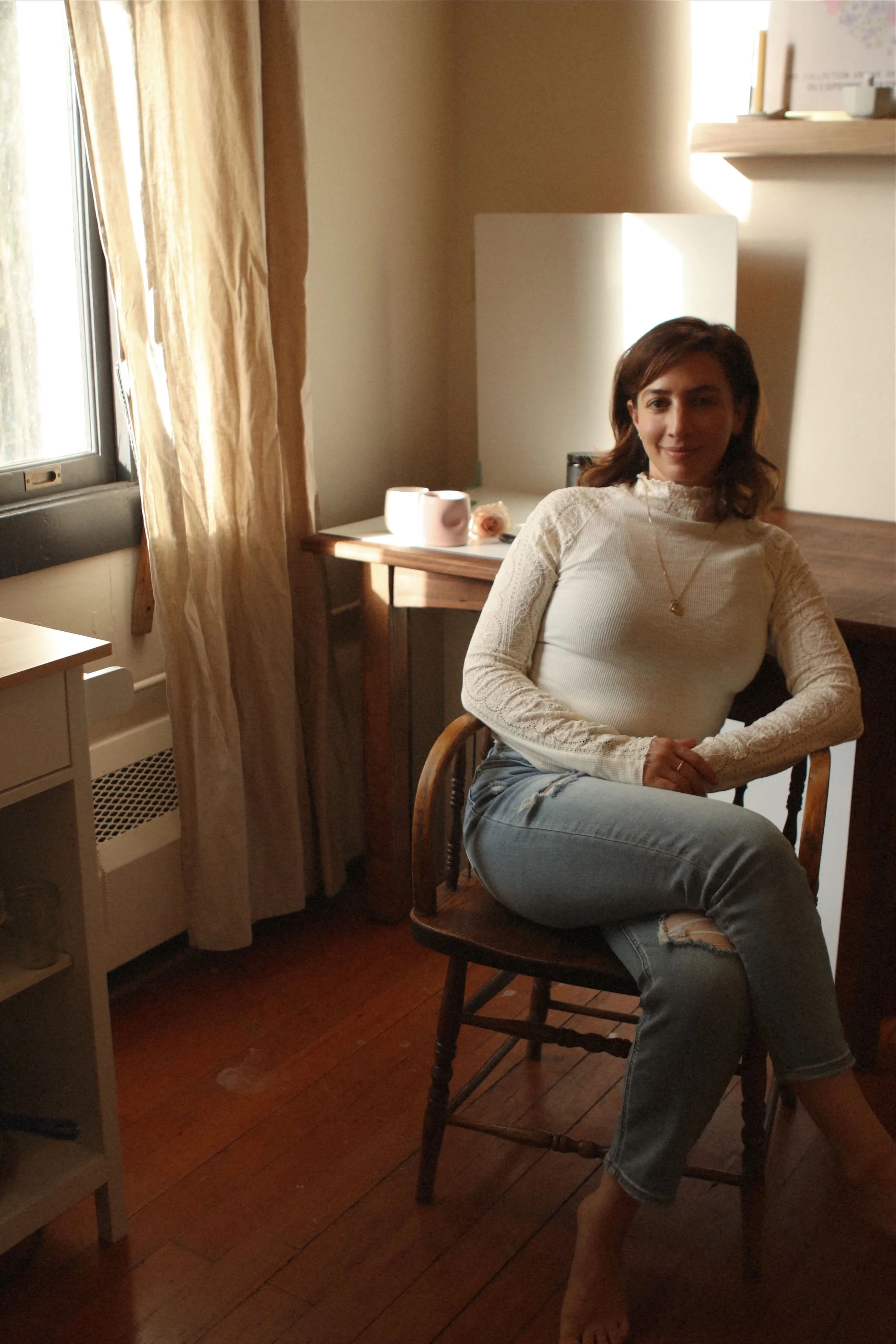 A woman sitting on a wooden chair in a warmly lit room, with a window and curtains on the left, a lamp and decorative items on the table behind her, and a shelf on the wall.