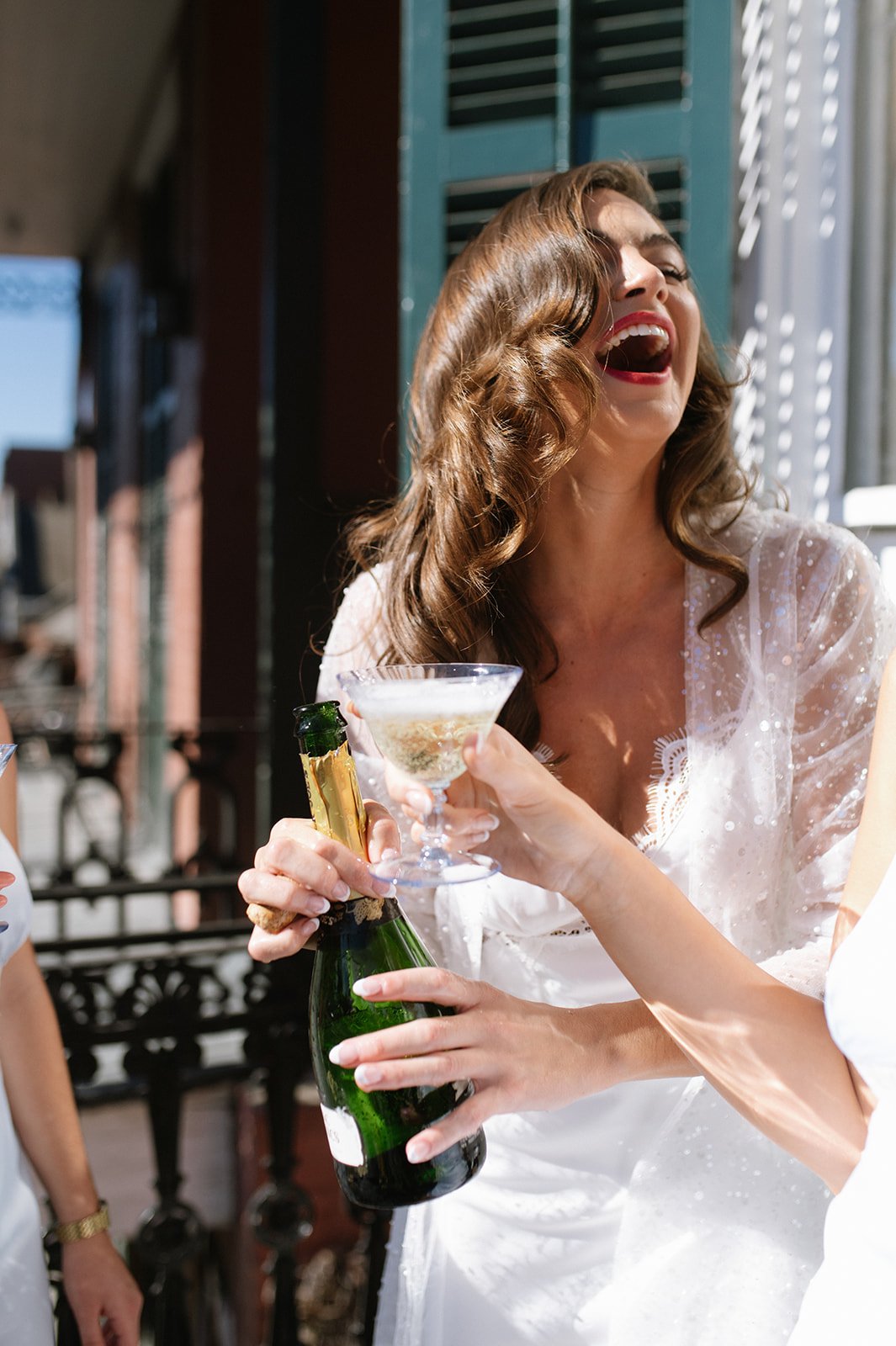 Woman with brown hair and red lipstick laughing, holding a champagne bottle and glass, during a celebration outside.