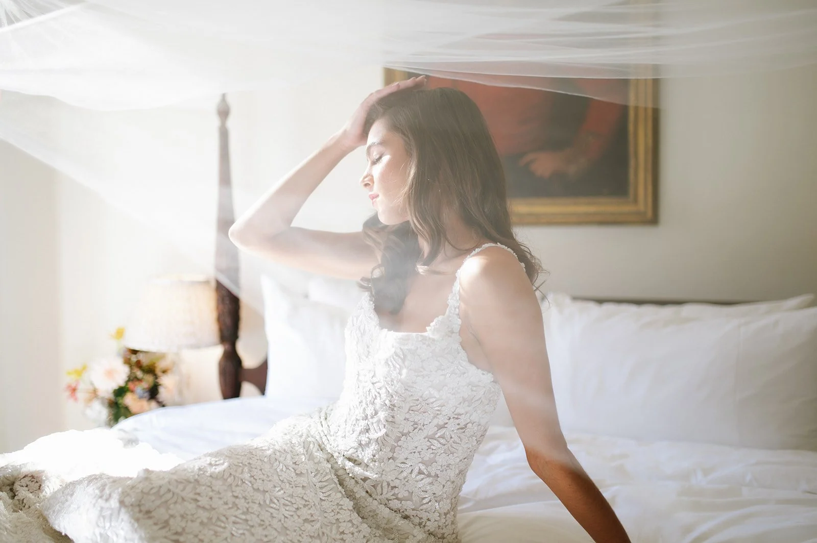 A woman in a white lace dress sitting on a bed in a softly lit room, with her hand on her head, eyes closed, and a painting on the wall behind her.