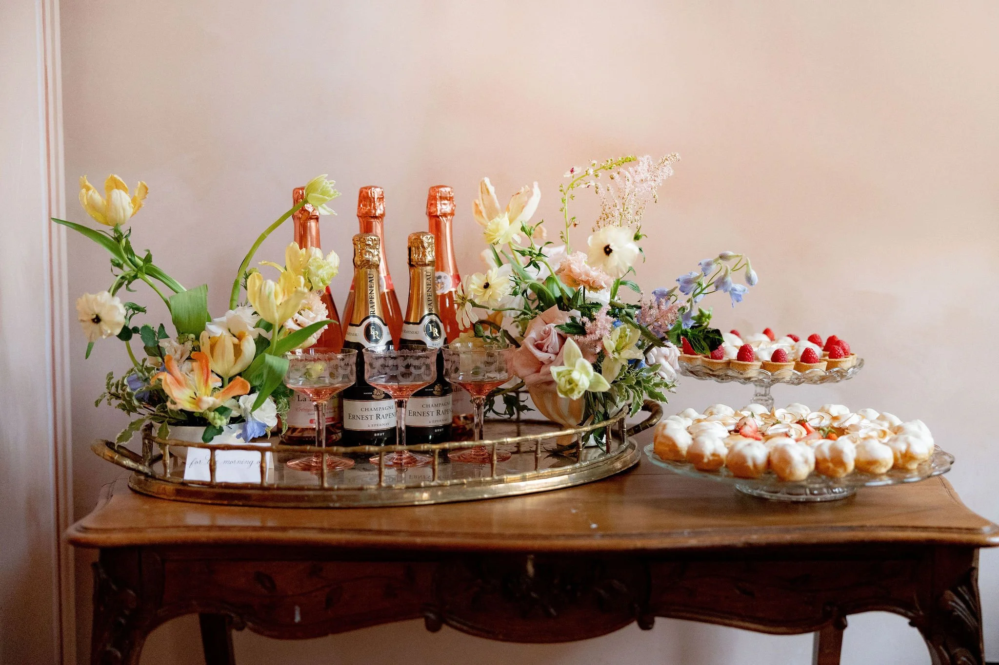 A wooden table with a silver tray holding four bottles of rosé champagne, glasses of champagne, and floral arrangements with white, pink, purple, and yellow flowers. To the right, there are two glass dessert trays filled with small, round pastries to