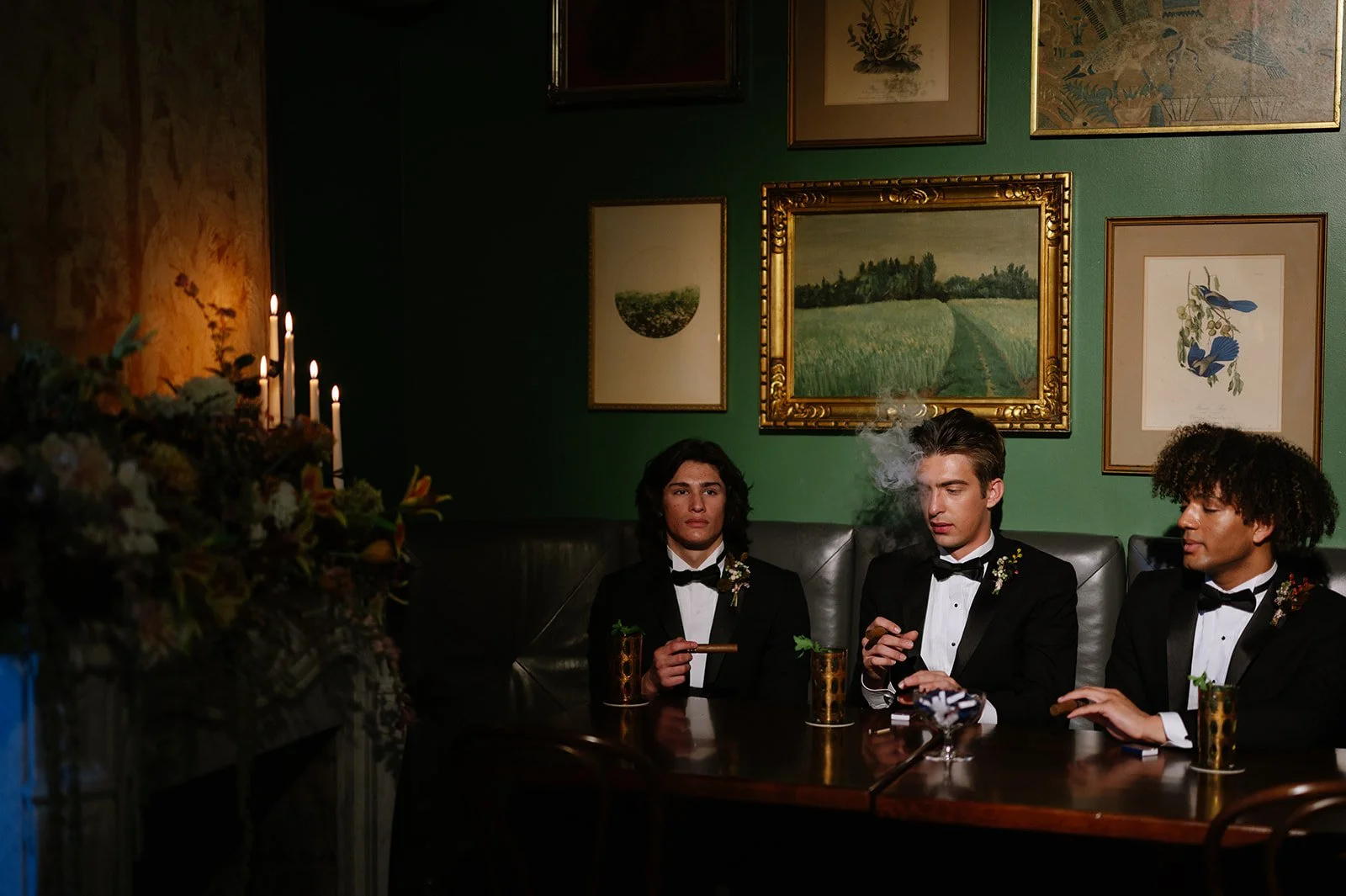 Three men in tuxedos sit at a dining table, smoking cigars in a dimly lit room with green walls and framed artwork.