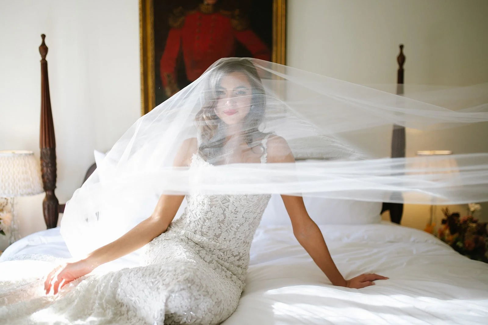 A woman in a wedding dress with a veil sitting on a bed in a room with a portrait and lamps.