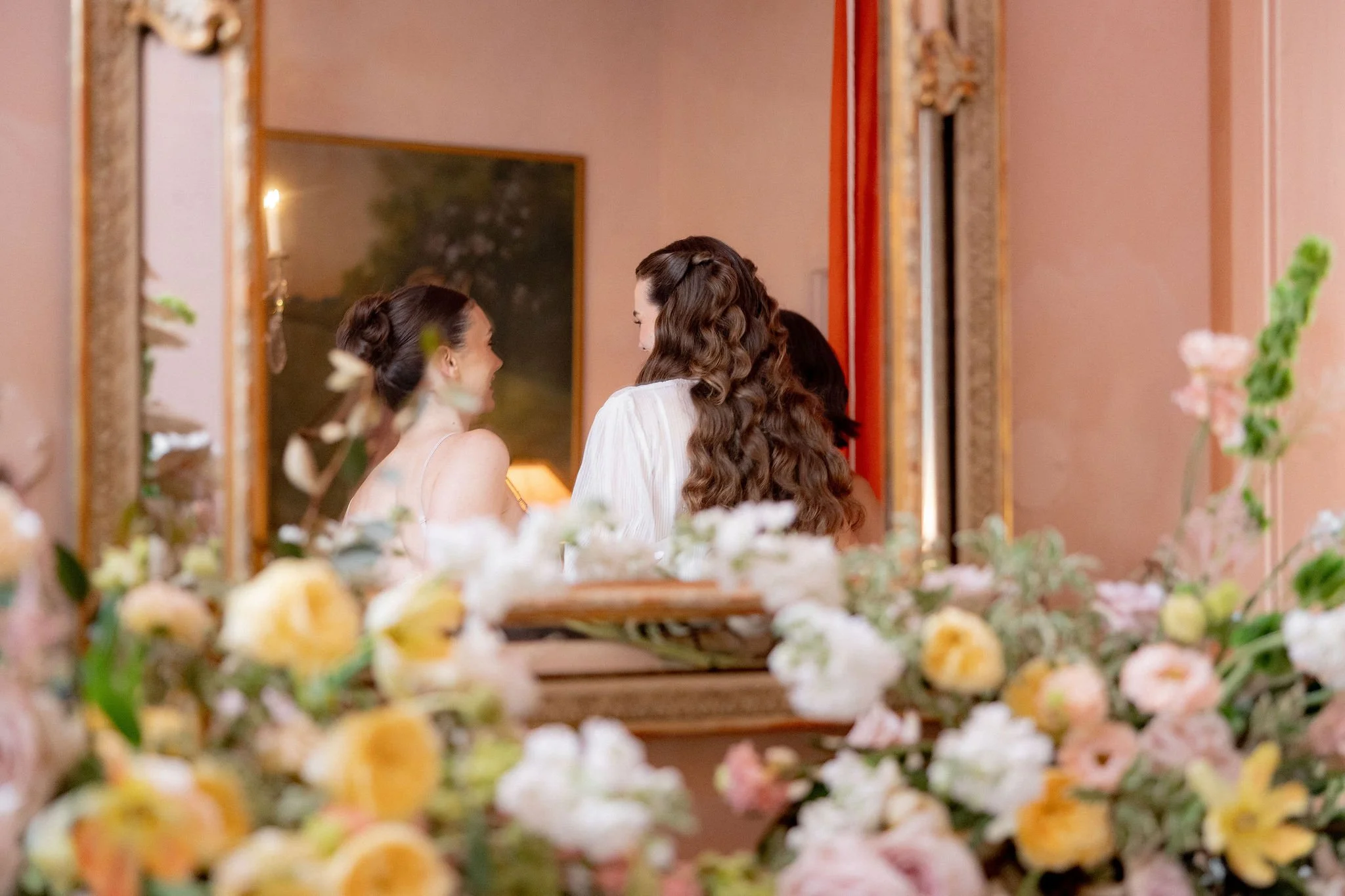 Two women with long, wavy hair, one with dark brown hair, are seen looking at each other through a mirror in a room decorated with pink walls and floral arrangements, with their reflections visible.
