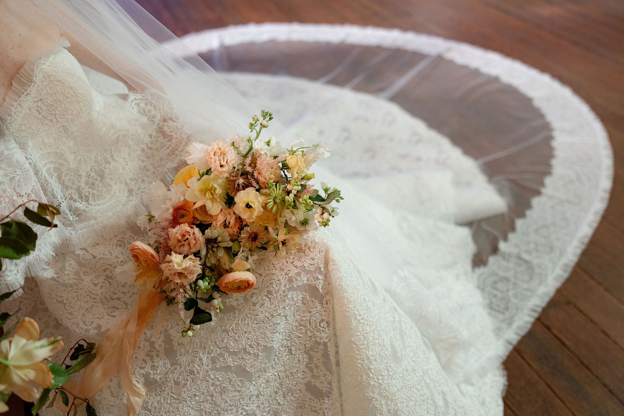 Wedding bouquet of pastel flowers placed on lace wedding dress that is spread on a wooden floor.