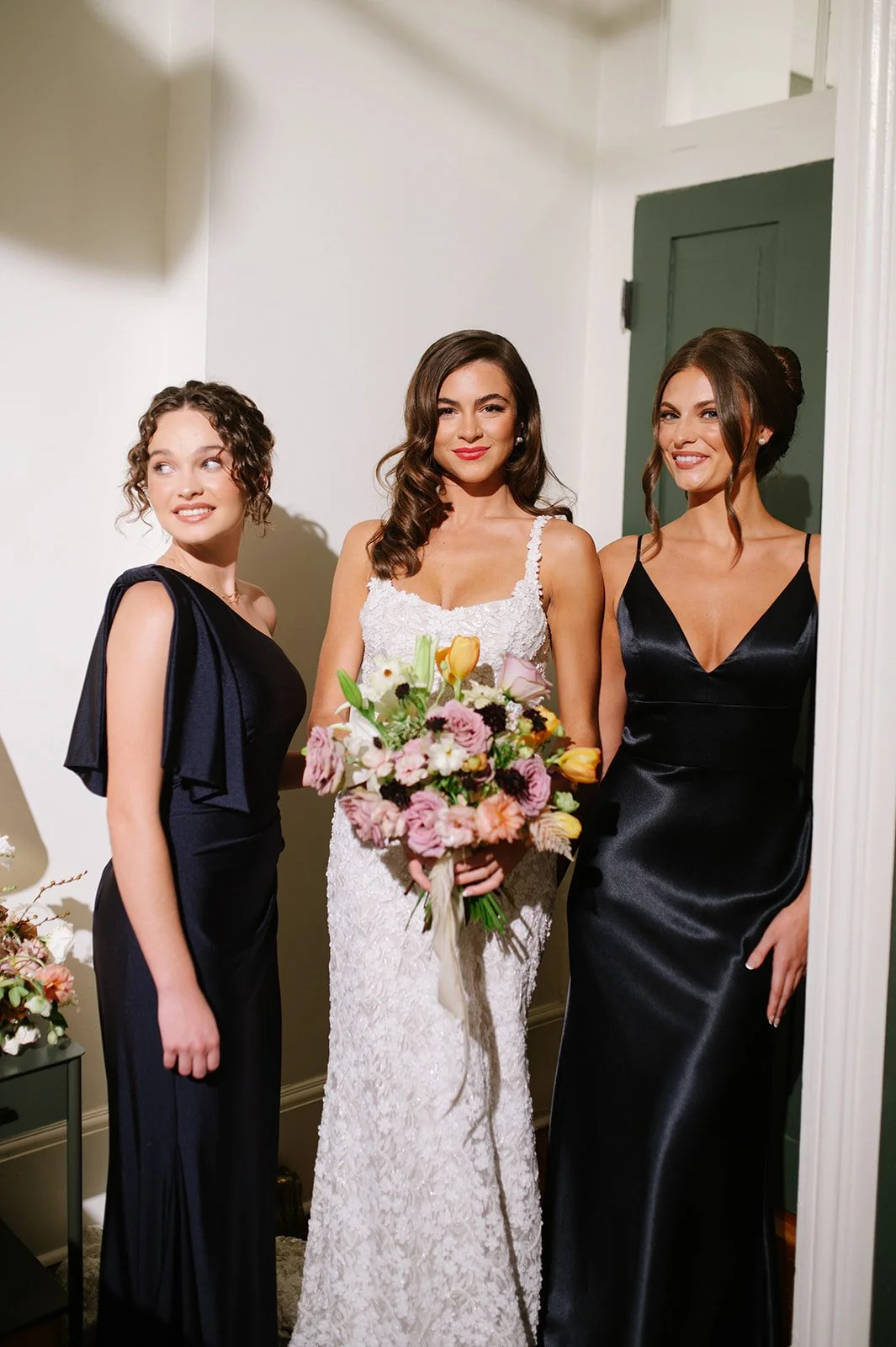Three women in formal dresses, with the woman in the center holding a bouquet of flowers, posing indoors.
