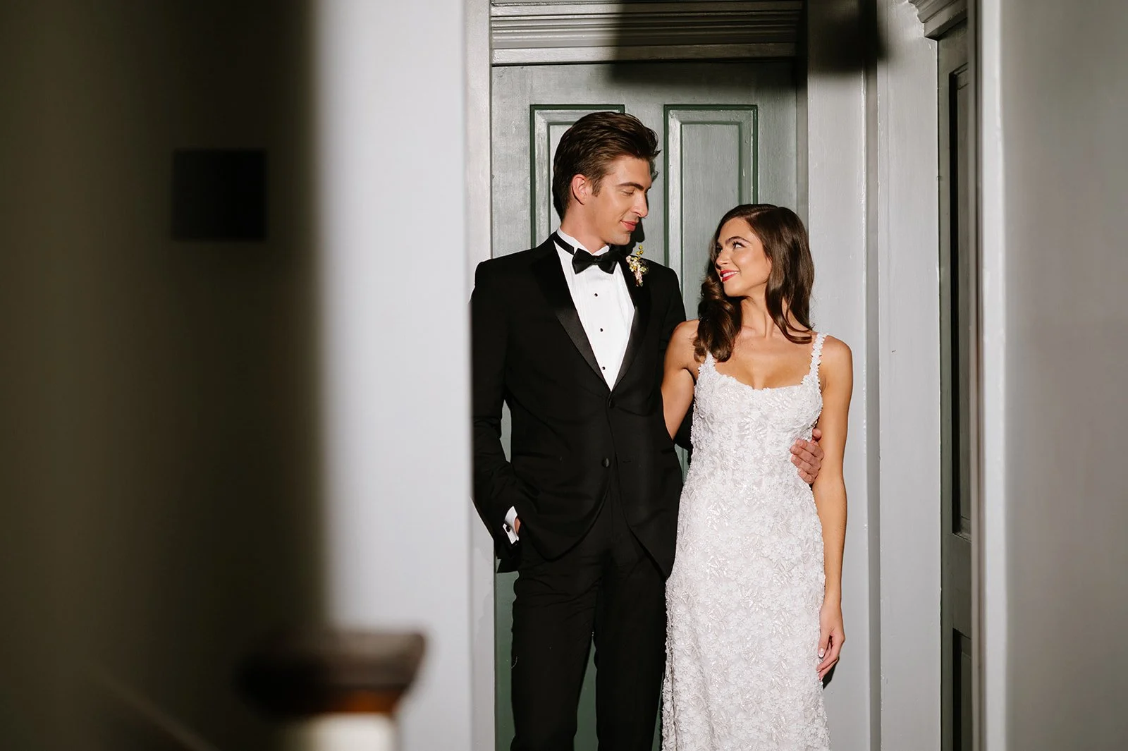 A bride and groom stand close together, gazing into each other's eyes, inside a house with a gray door in the background. The groom wears a black tuxedo with a bow tie, and the bride wears a white lace wedding gown. They are smiling warmly at each ot