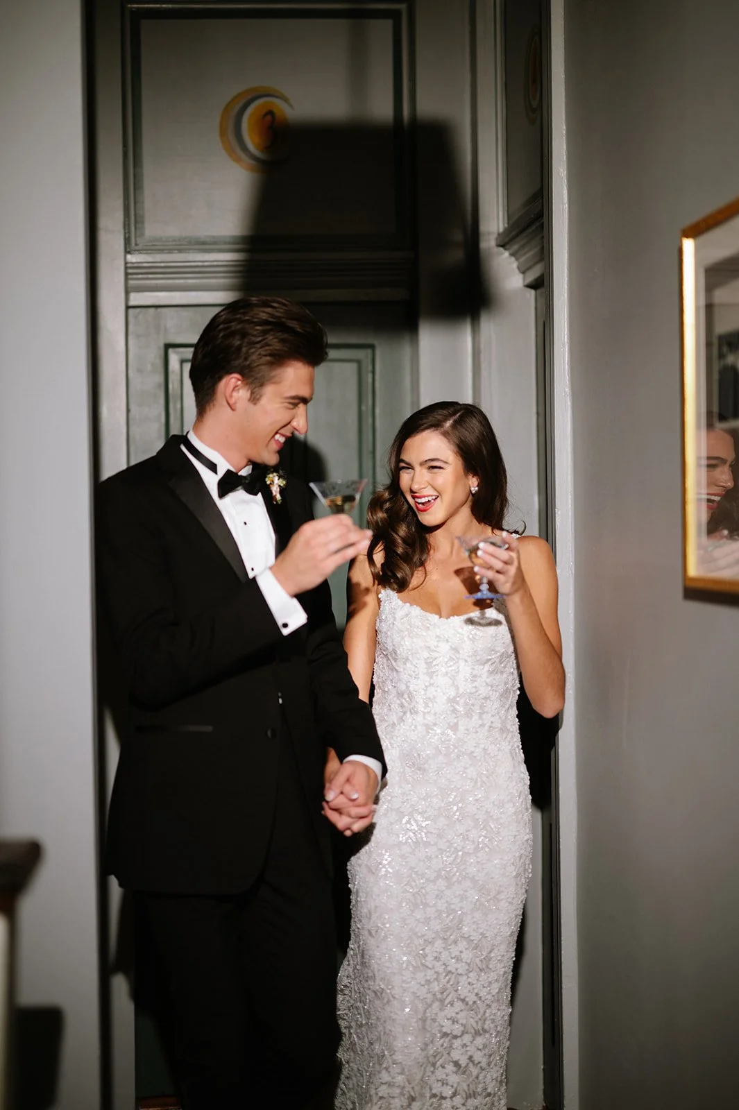 A newlywed couple, the groom in a black tuxedo and the bride in a white lace wedding dress, sharing a joyful moment and holding drinks during their wedding.
