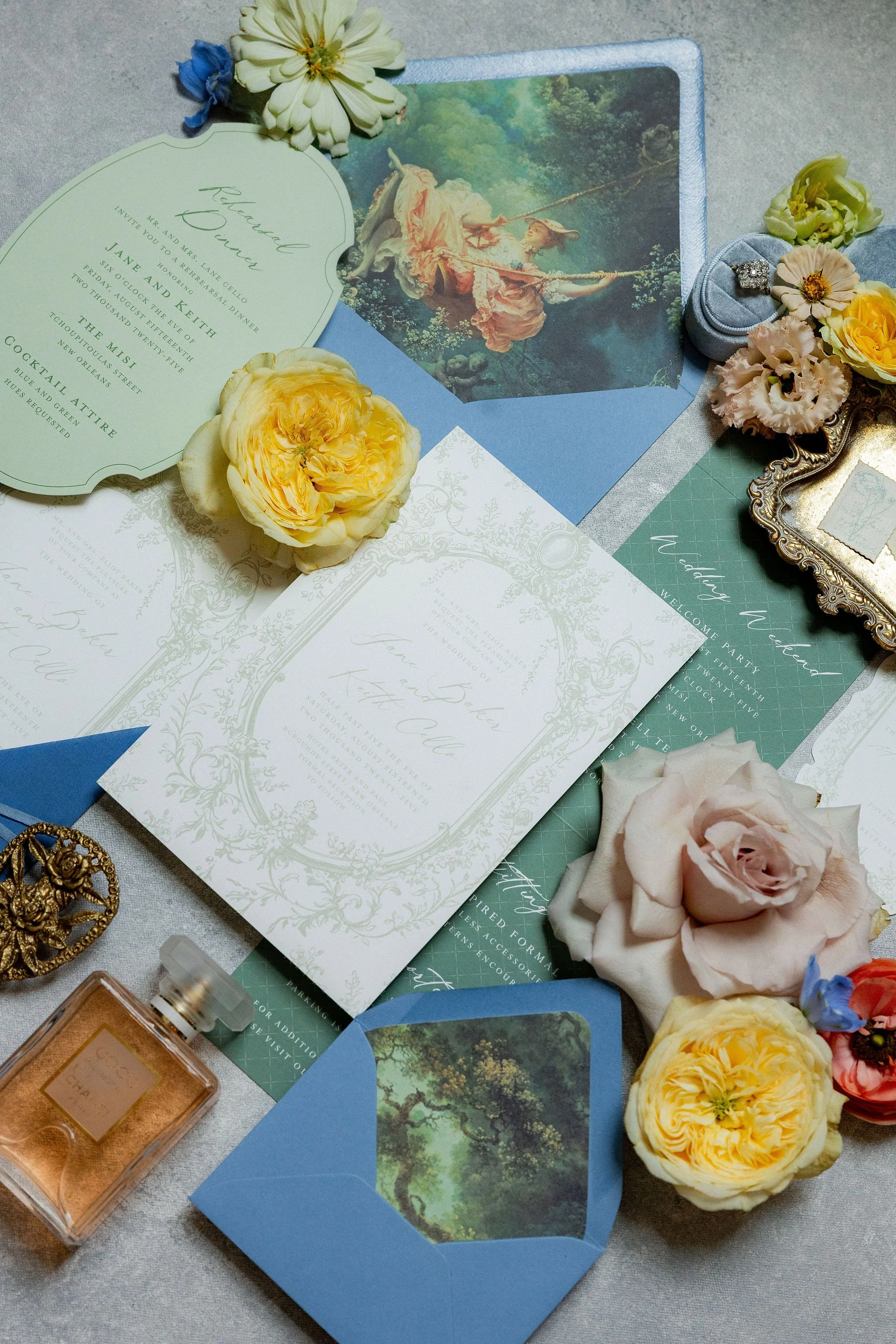 A wedding flat lay featuring floral arrangements, wedding invitations, a perfume bottle, a ring box with rings, and decorative items on a grey surface.
