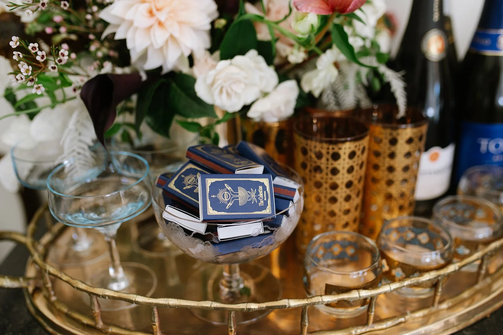 A decorative tray holding several empty glasses, a bowl filled with matchboxes labeled 'The Crystal,' two amber-patterned glasses, and bottles, with a flower arrangement of white and pink flowers in the background.