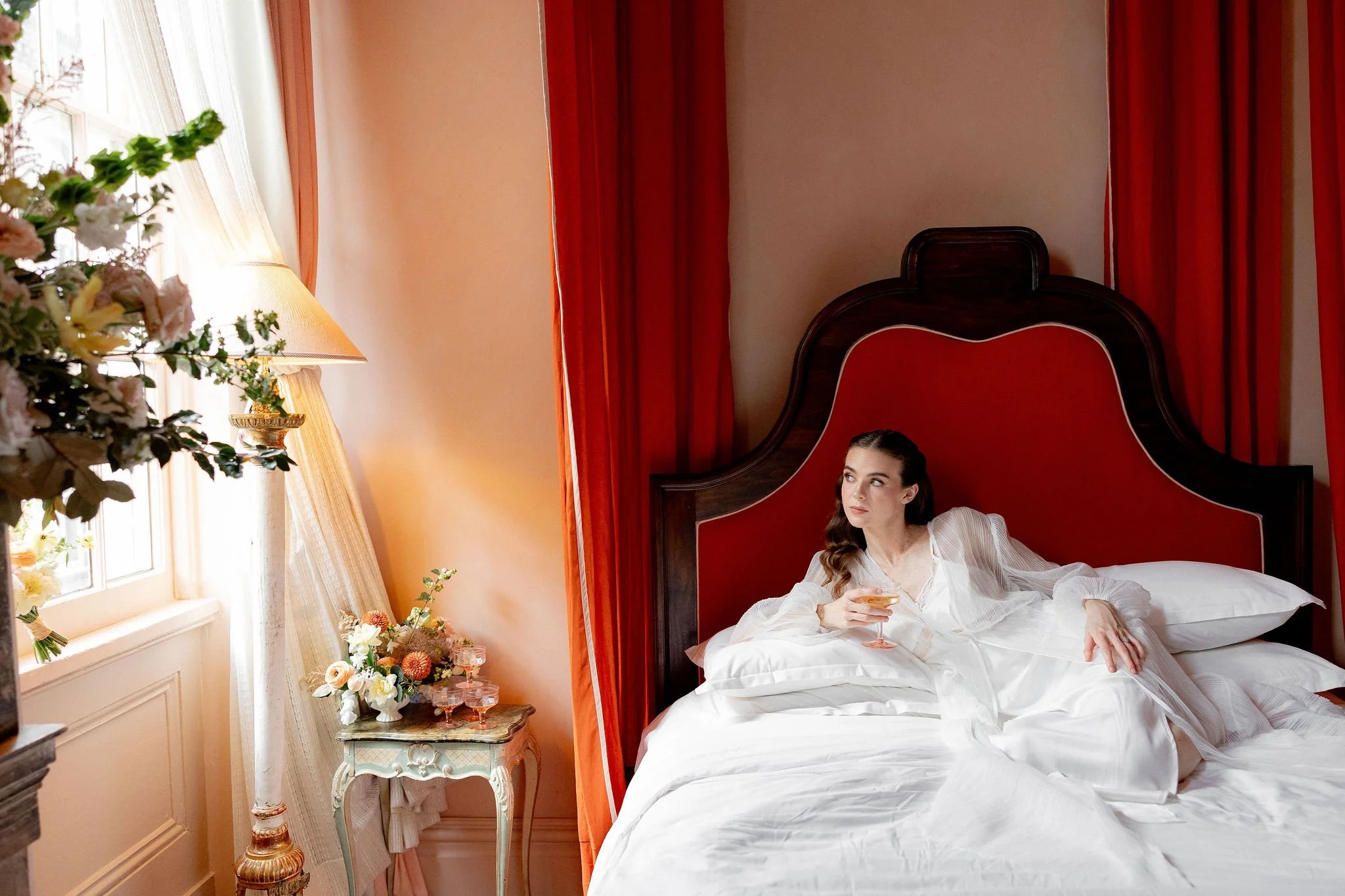 Woman in white pajamas relaxing on a bed in a decorated bedroom with red curtains, a floral arrangement, and a side table with a lamp and flowers.