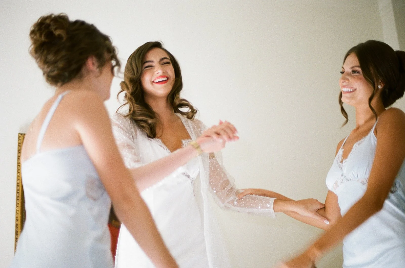 Three women in white dresses laughing and smiling together in a bedroom.