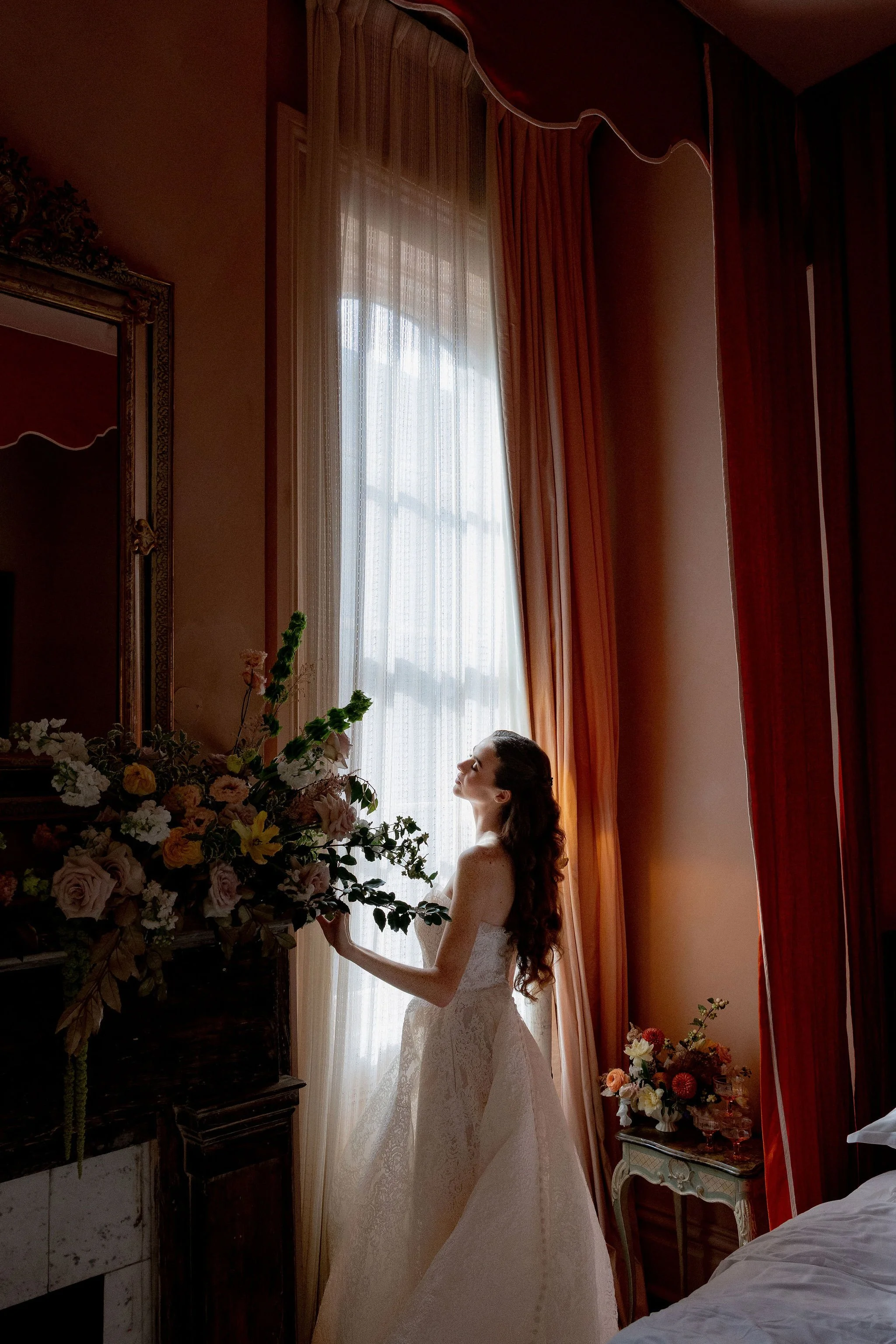 A woman in a wedding dress stands by a large window with sheer curtains, holding a bouquet of flowers, in a warmly lit room with orange curtains and floral arrangements.