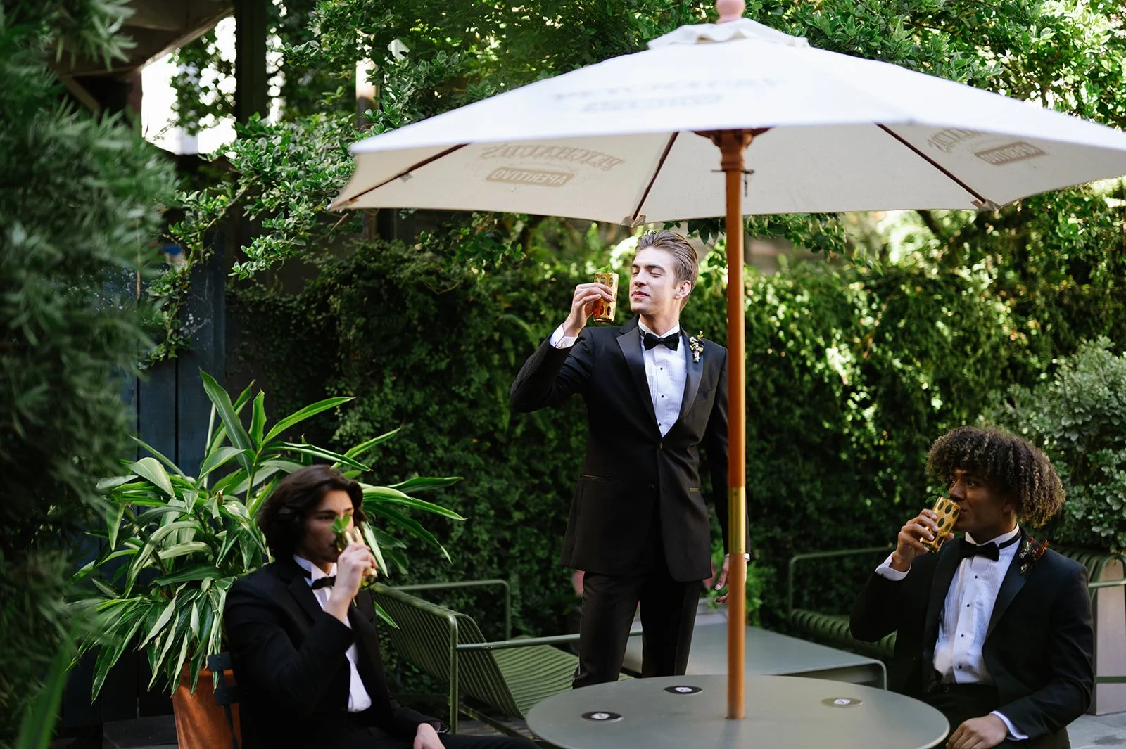 Three young men in tuxedos enjoying drinks outdoors under a large umbrella at a lush garden setting.