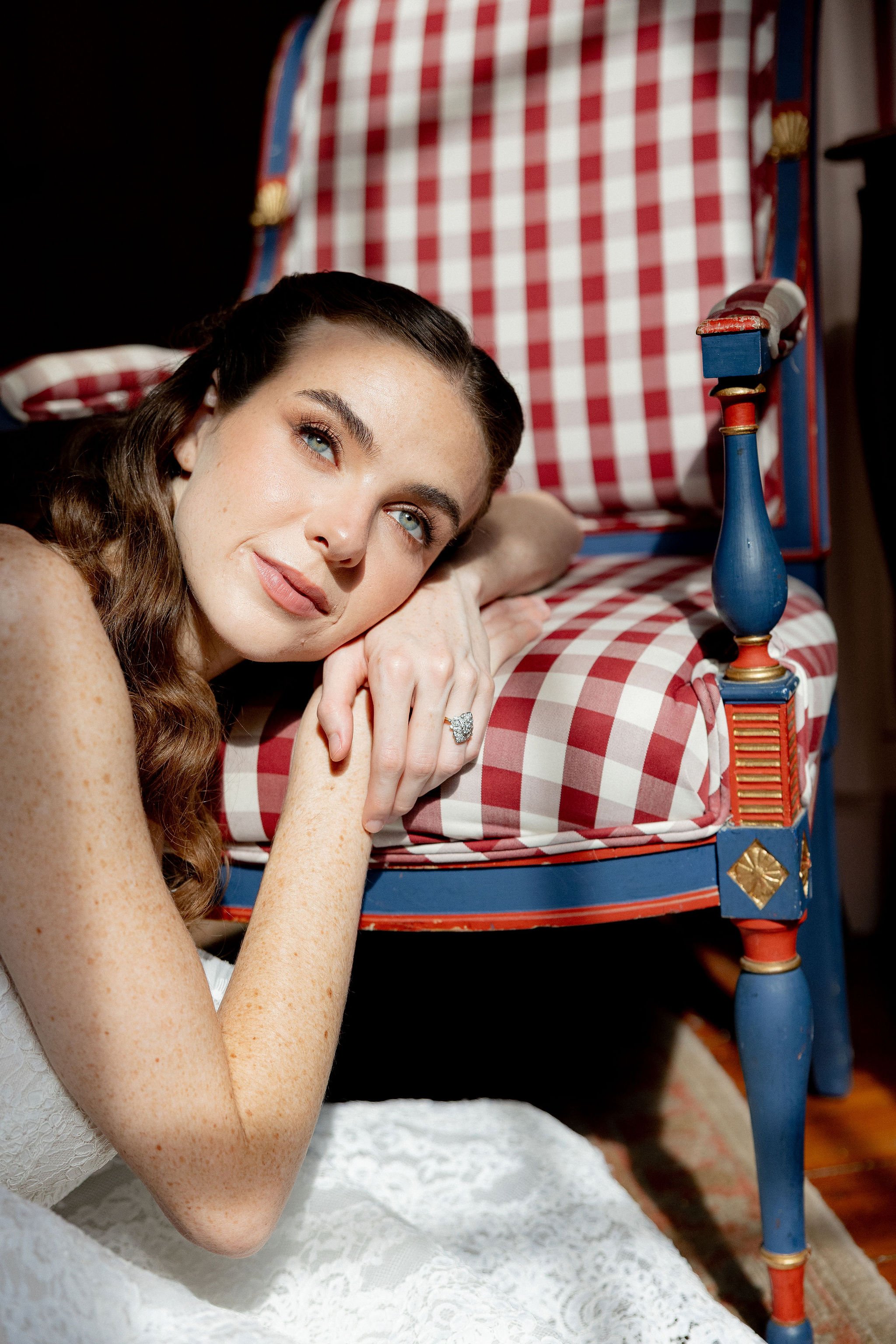 A young woman with curly brown hair and blue eyes lying beside a vintage red, white, and blue checkered bed. She is resting her head on her arm and looking ahead, wearing a white lace dress.