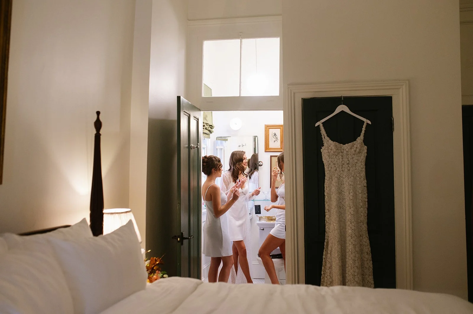 Four women in white robes are inside a small room, preparing and talking to each other. A wedding dress is hanging on a black door to the right.
