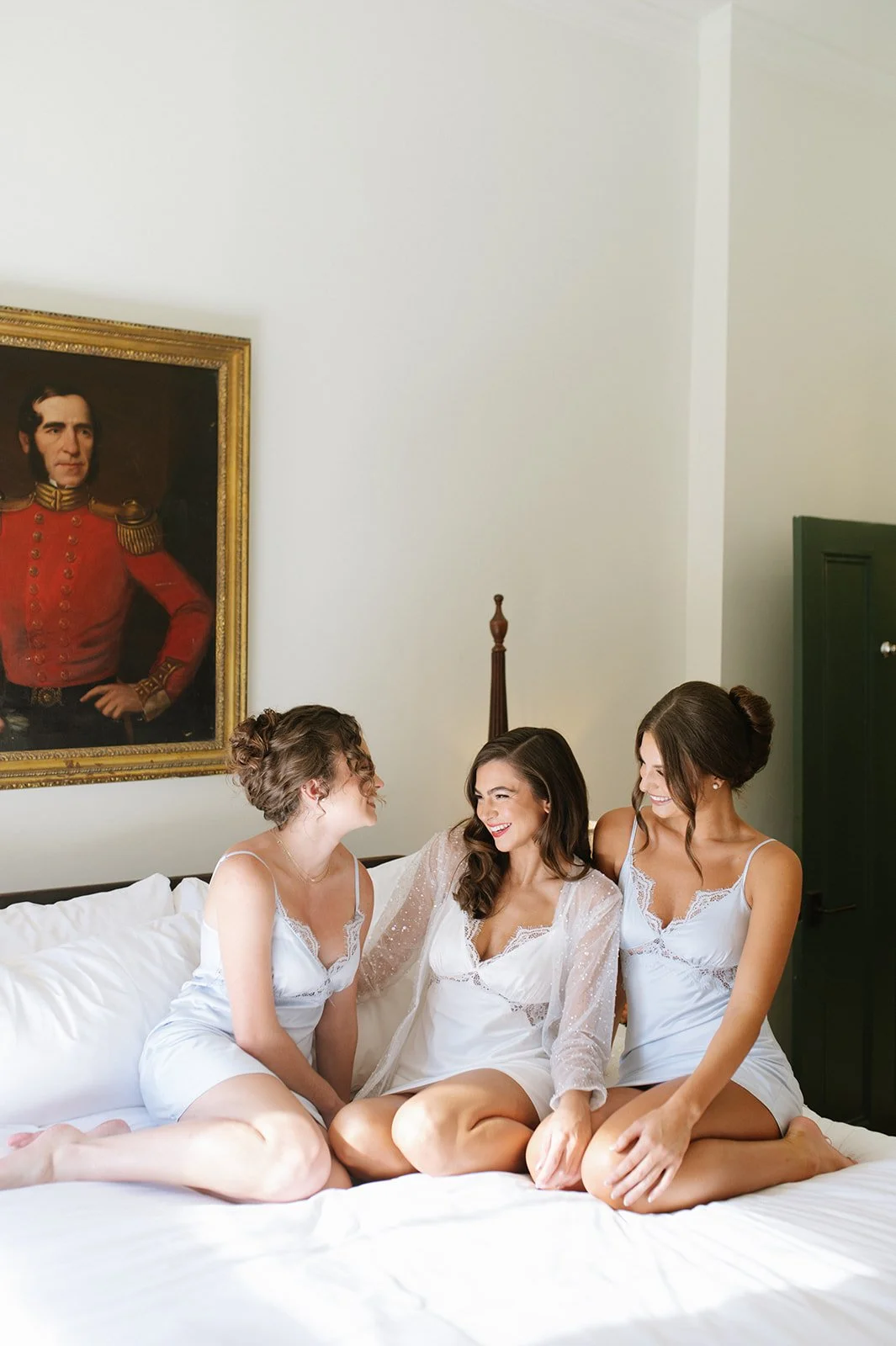 Three women in white nightgowns sitting on a bed, smiling and talking. Behind them is a framed portrait of a man in a red military uniform.