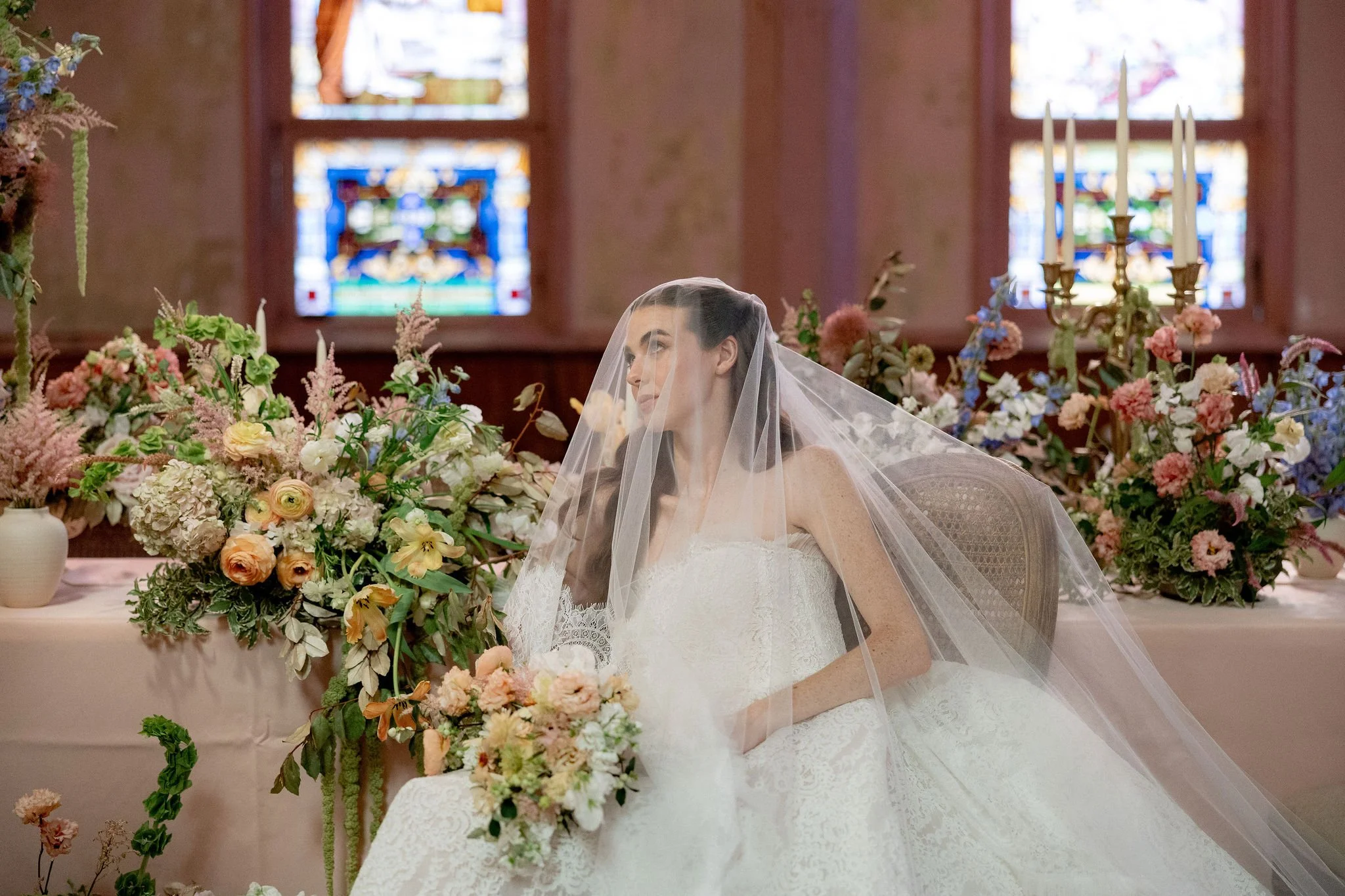 A bride in a wedding dress with a veil sitting at a table decorated with a large bouquet of pastel-colored flowers and surrounded by other floral arrangements, inside a church with stained glass windows.