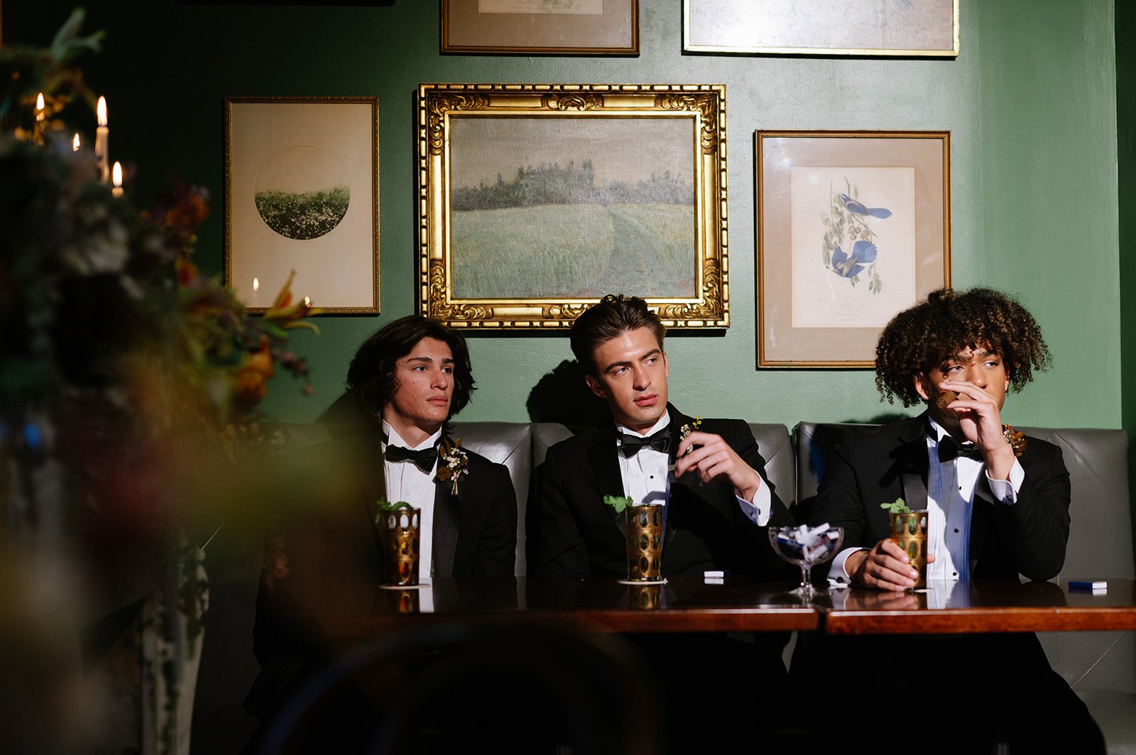 Three young men dressed in tuxedos sitting at a table in a restaurant or lounge, with green walls and framed artwork behind them. One is holding a drink, another has his hand near his mouth, and the third is looking straight ahead with a serious expr