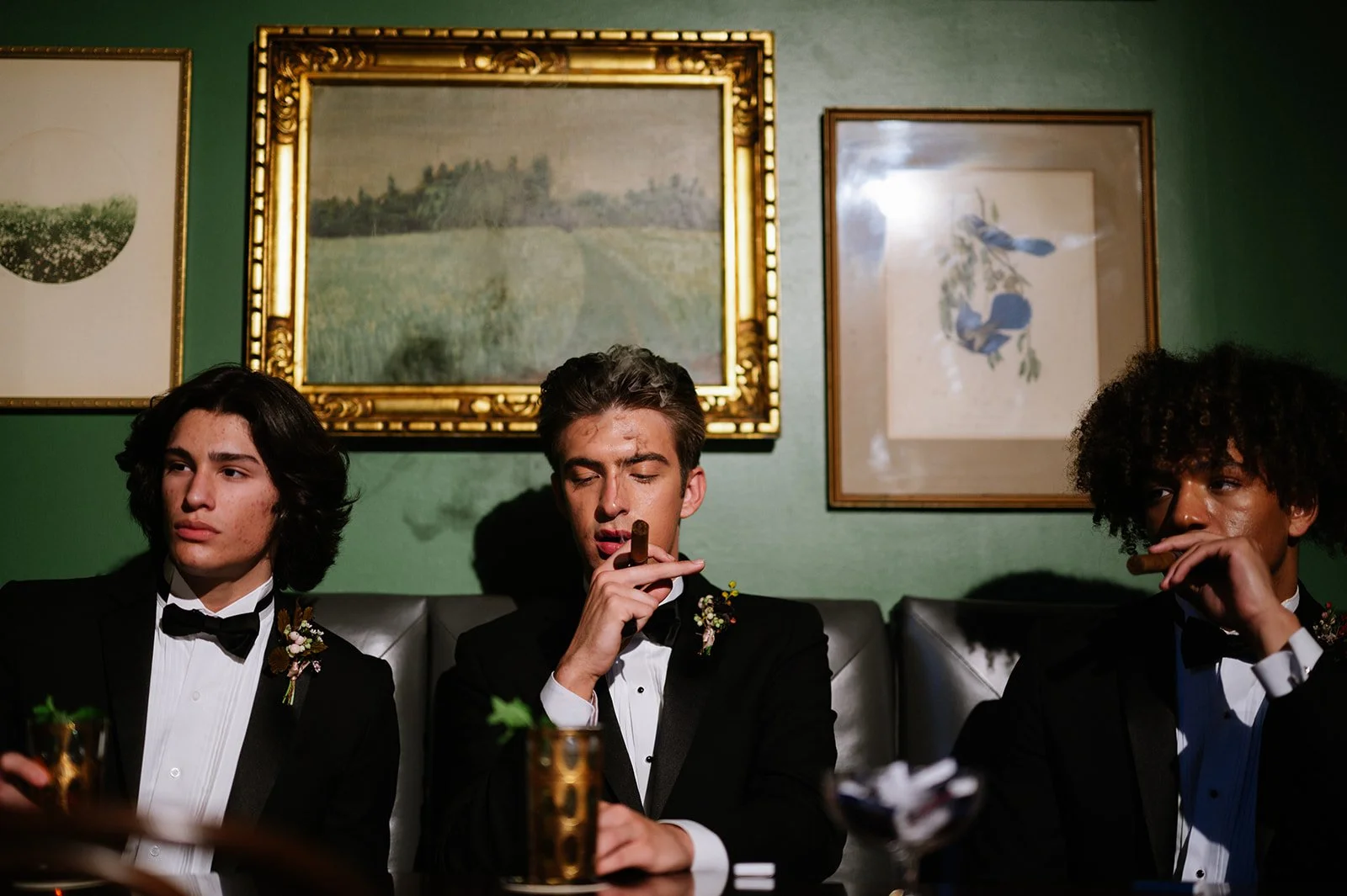 Three young men in tuxedos sitting at a table, two smoking cigars, in an art-filled room with framed paintings on green walls.