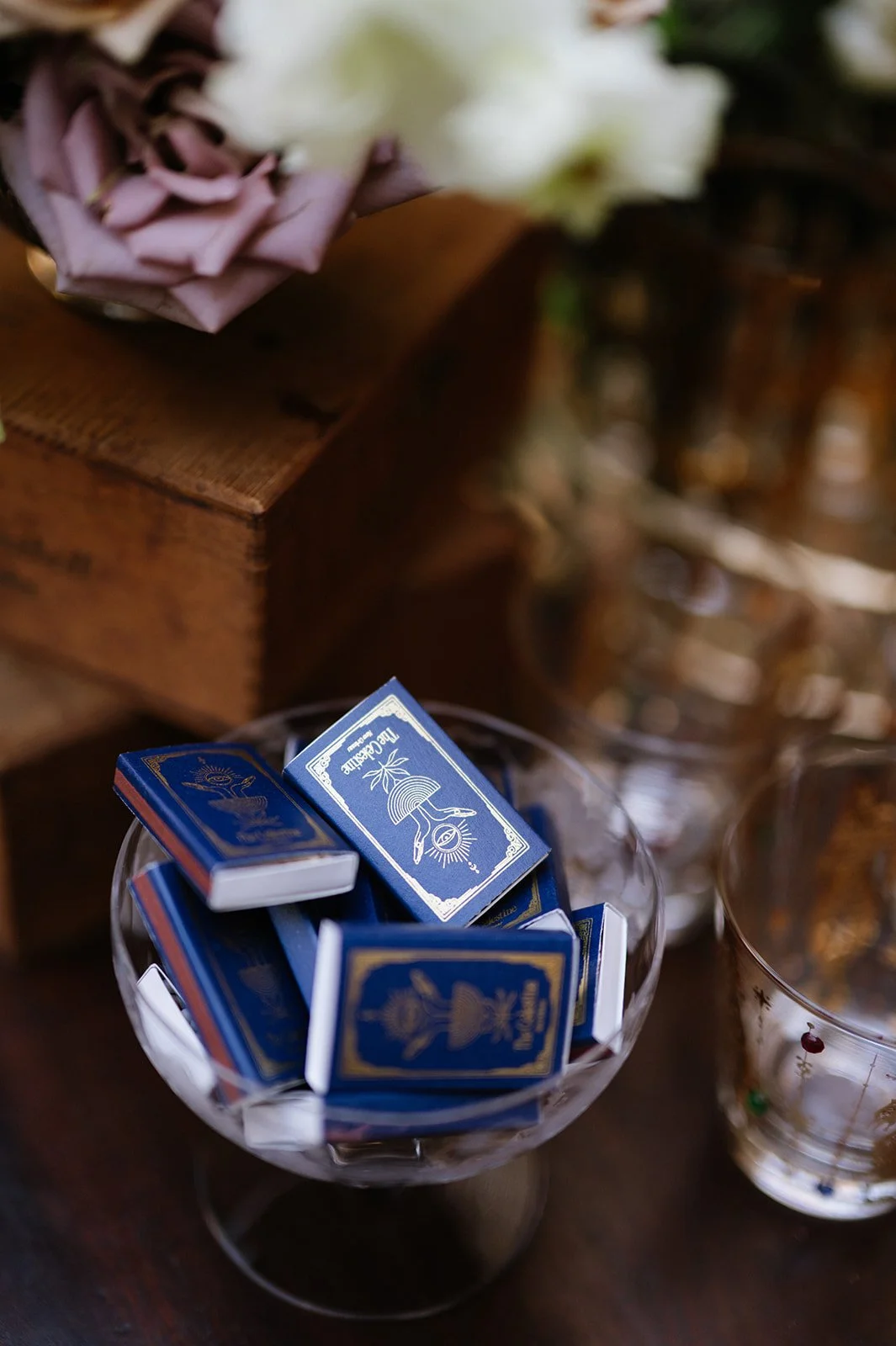 Blue and white matchboxes in a glass dish on a wooden table, with a partially visible floral arrangement in the background.