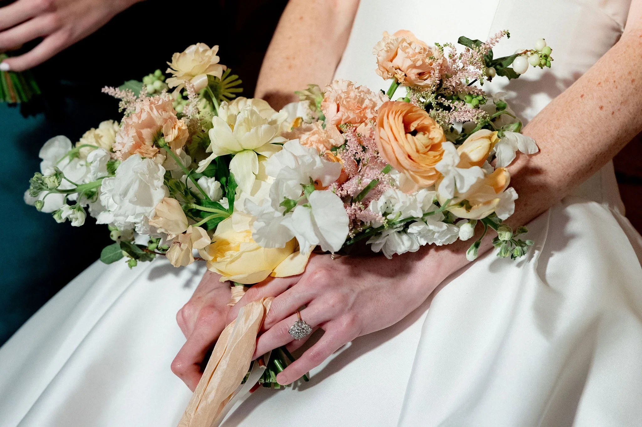 Person holding a bridal bouquet of peach, white, and pale pink flowers, with a wedding dress and a diamond ring visible.