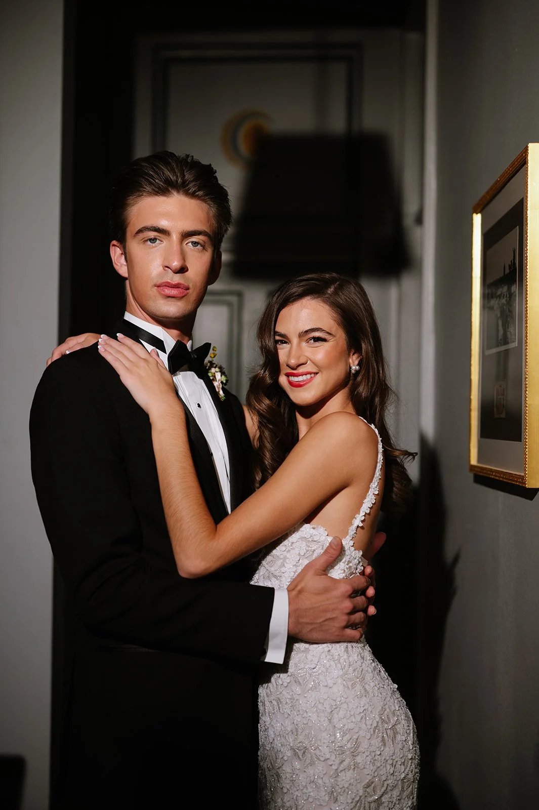 A bride and groom in wedding attire standing close together in an indoor setting with framed pictures on the walls.