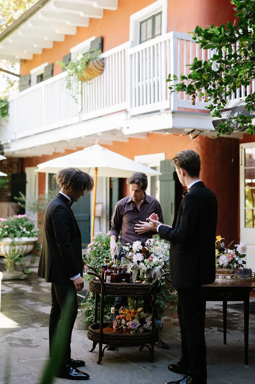 Three men in tuxedos are gathered around a table with bottles and flowers outside, having a conversation, in a backyard with a two-story red house and patio umbrellas.