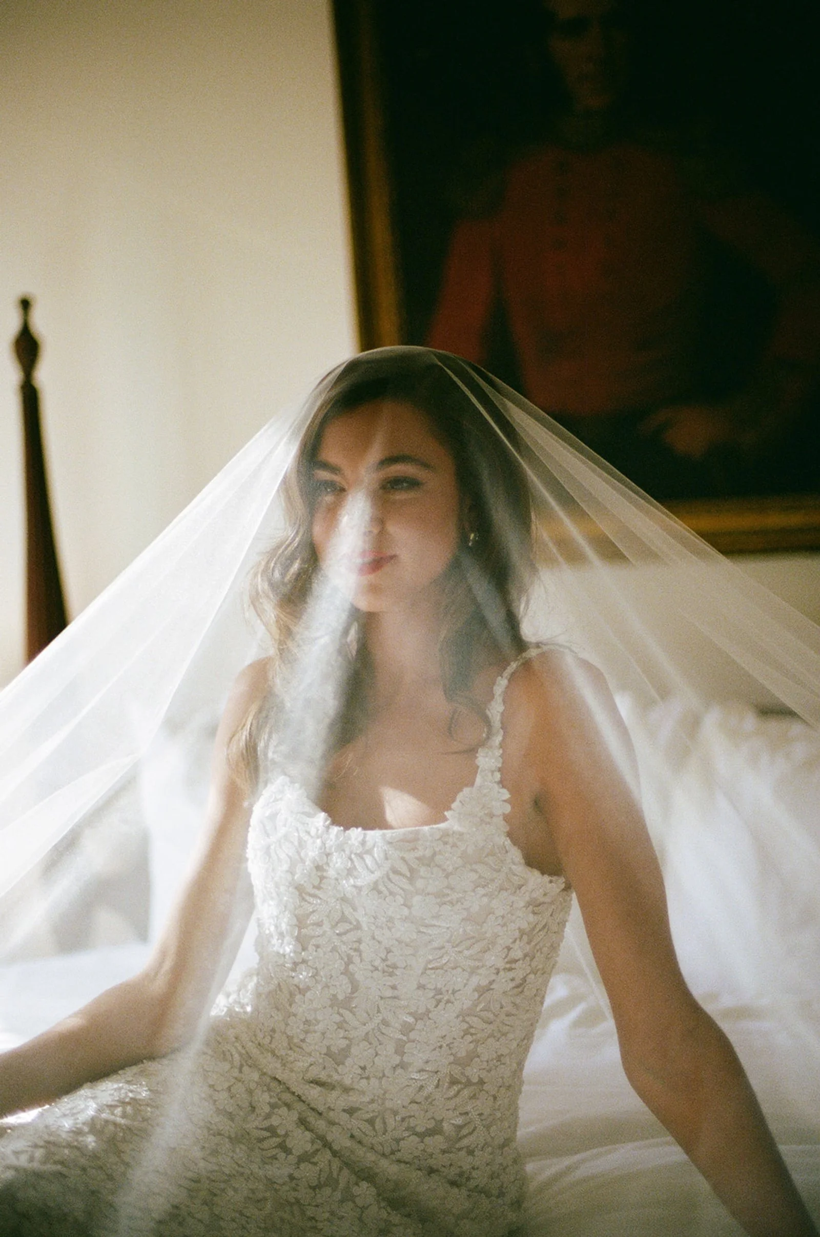 A bride with wavy brown hair, wearing a lace wedding dress and veil, sitting on a bed with soft natural light.