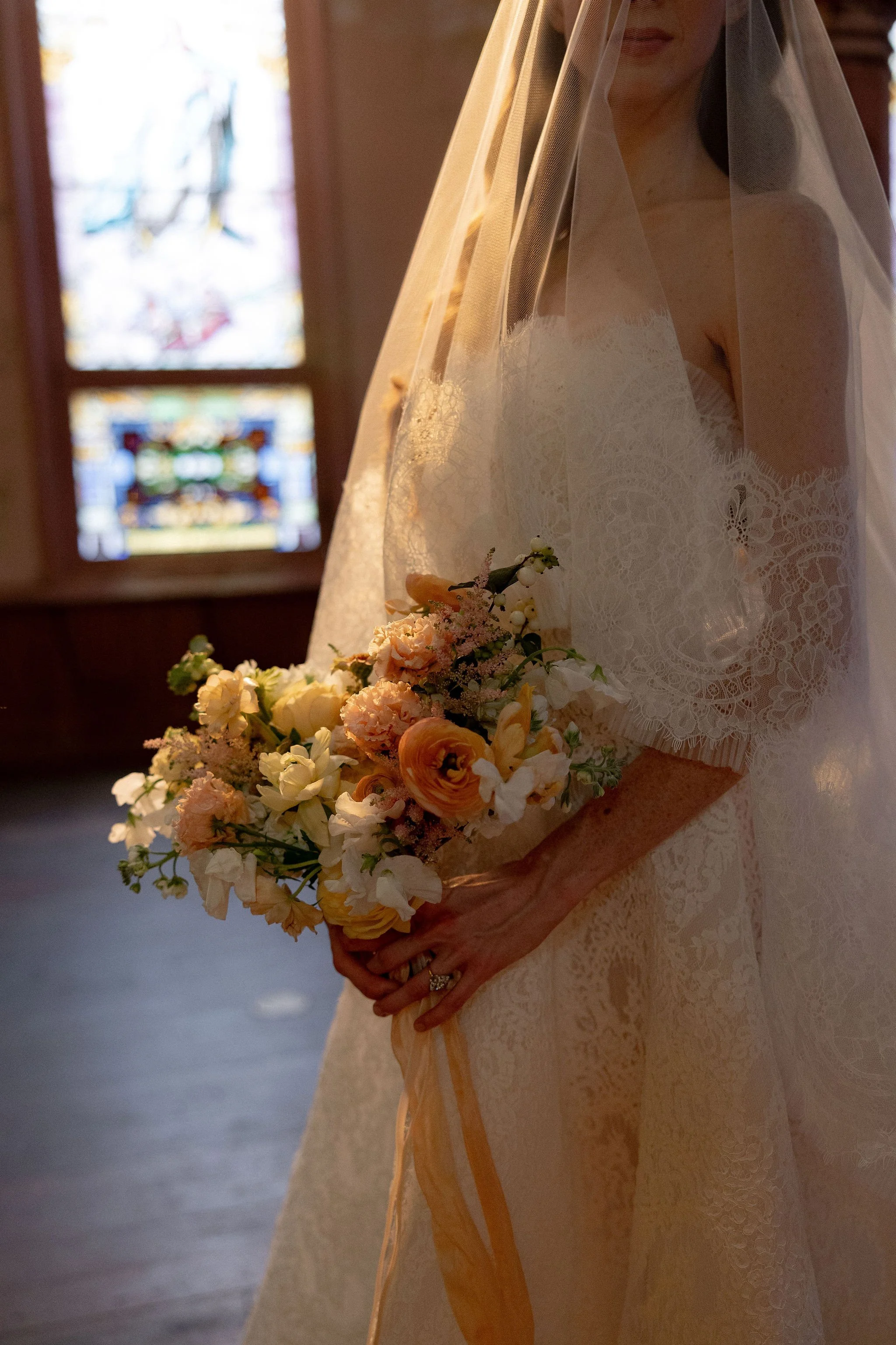 A bride in a lace wedding dress holding a bouquet of peach and white flowers, standing inside a church with stained glass windows in the background.