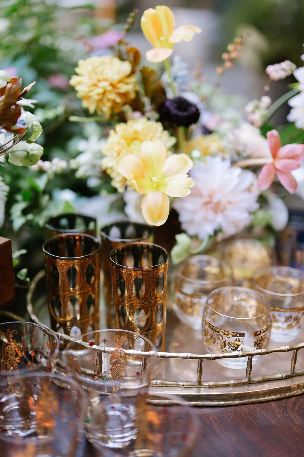 A decorative display featuring a large bouquet of various colorful flowers with pink, yellow, white, and black blooms, surrounded by several glass and metal cups with gold accents, arranged on a round tray on a wooden surface.