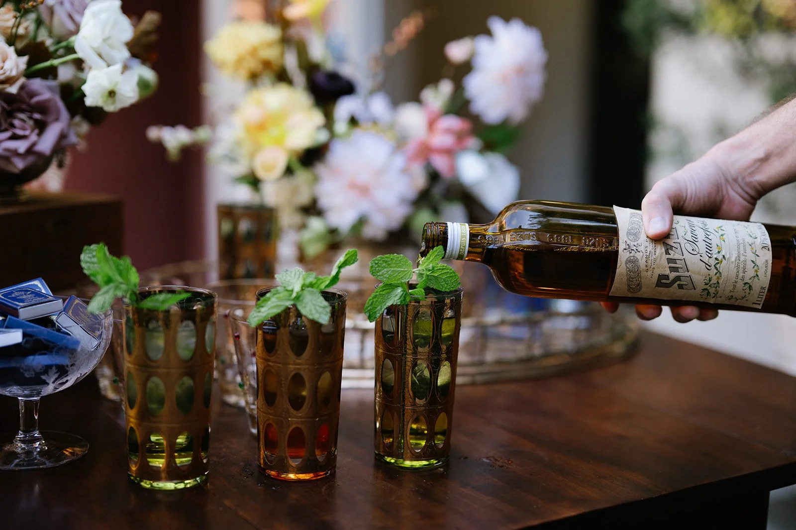 A hand pouring a dark liquid from a bottle into a glass with mint leaves, on a wooden table with colorful glasses and flowers in the background.