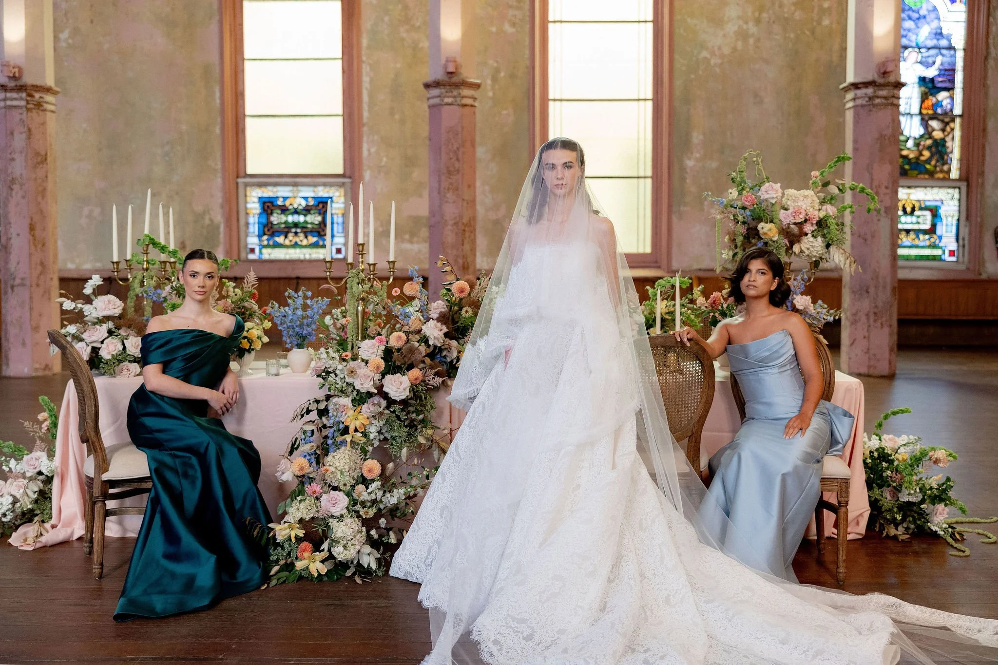 A bride in a wedding gown with a veil standing between two women in elegant dresses, seated at a decorated table with floral arrangements and candlesticks inside a church with stained glass windows.