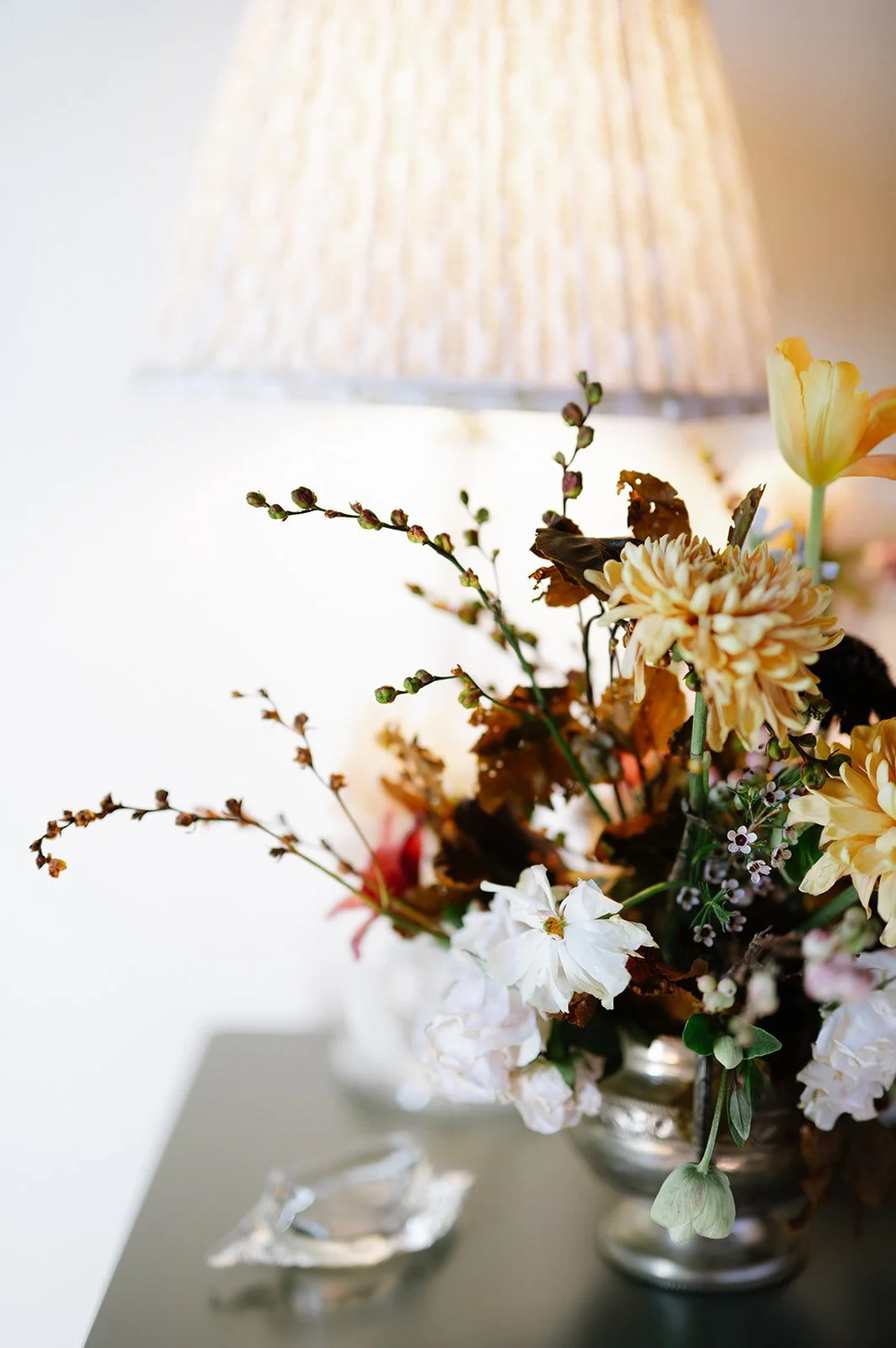 A floral arrangement with white, cream, and pale pink flowers in a silver vase, placed on a table next to a lit table lamp with a pleated lampshade.