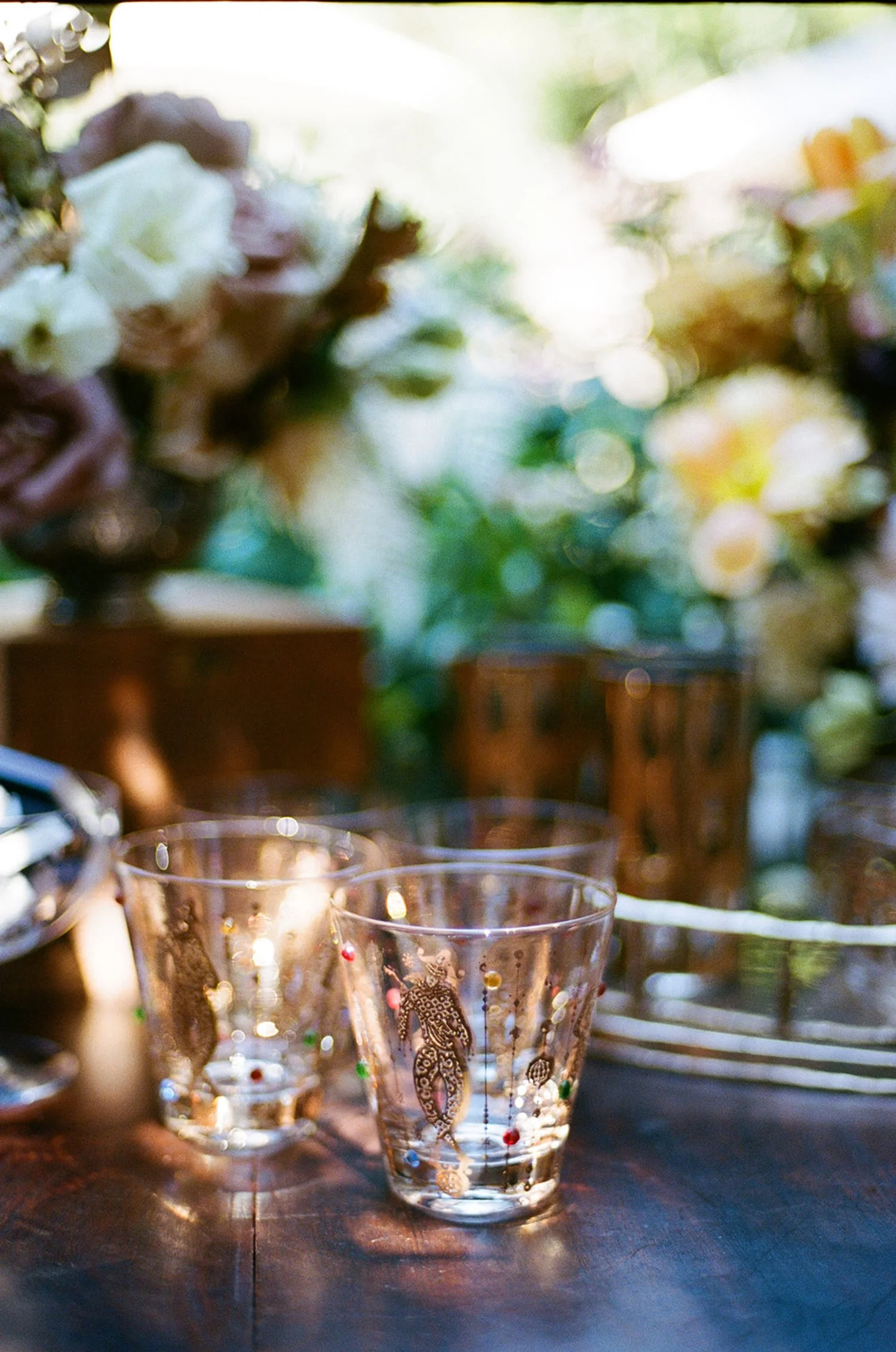 Glass cups with colorful decorative patterns on a wooden surface, with floral arrangements in the background