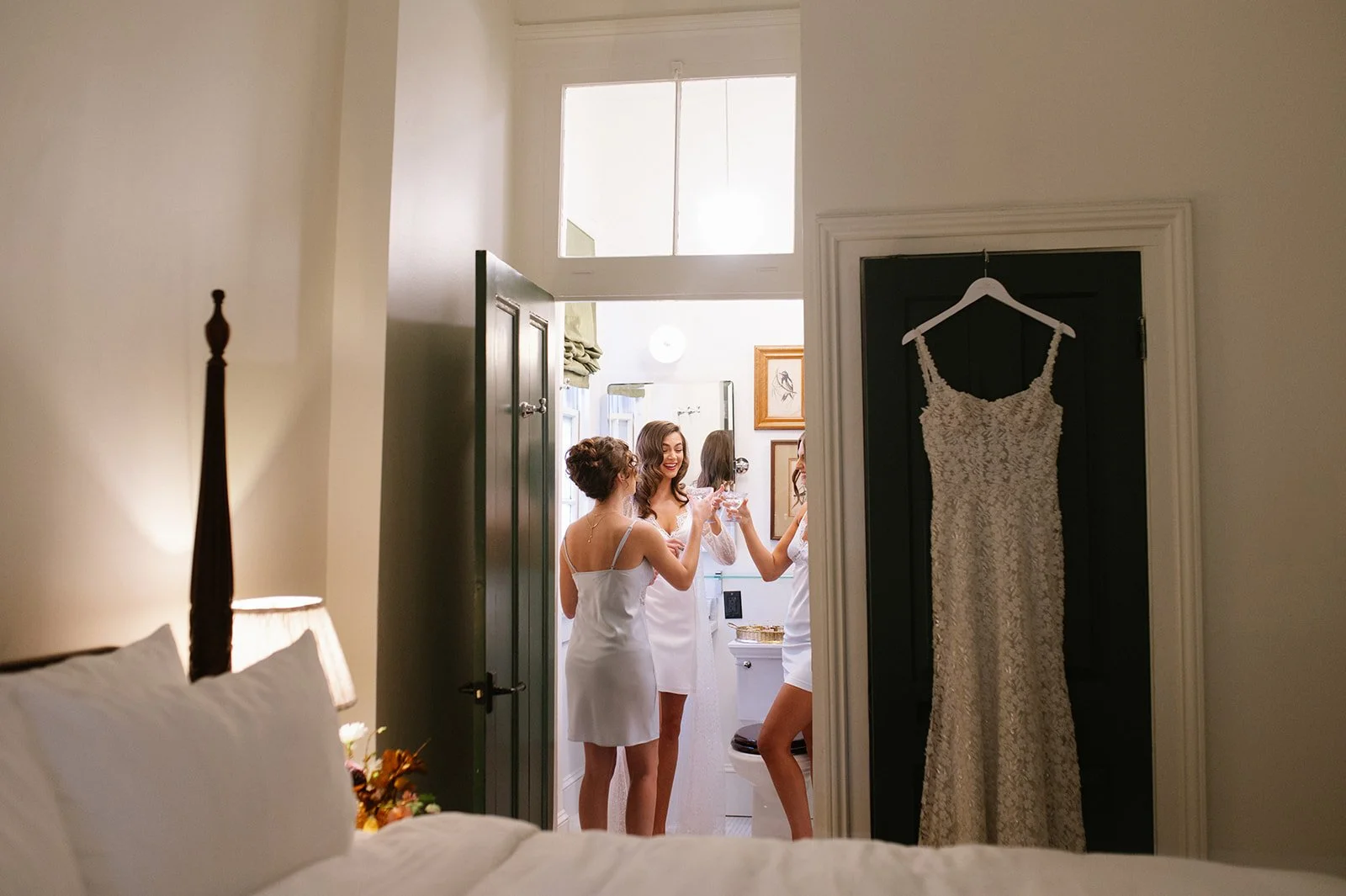 Three women in white dresses celebrating and toasting with glasses in a small bathroom, seen from a bedroom with a bed and a lamp in the foreground. A wedding dress is hanging on a door next to the bathroom.
