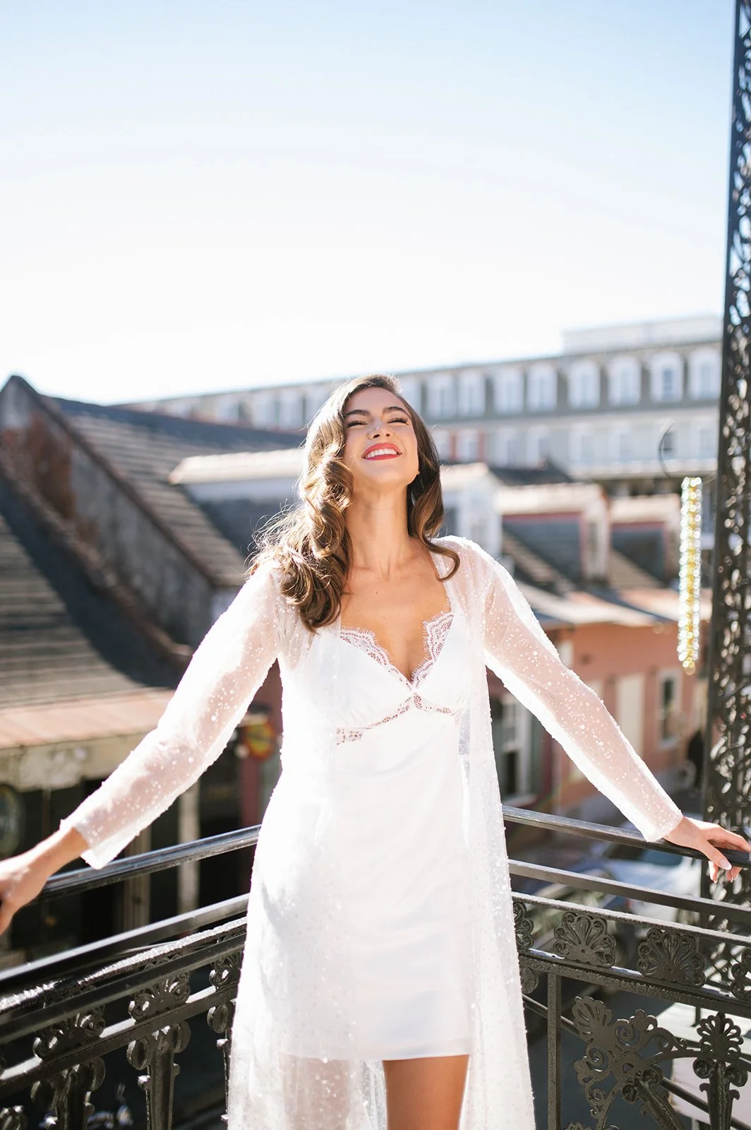 A woman in a white dress standing on a balcony with sunlight shining behind her, smiling with closed eyes.