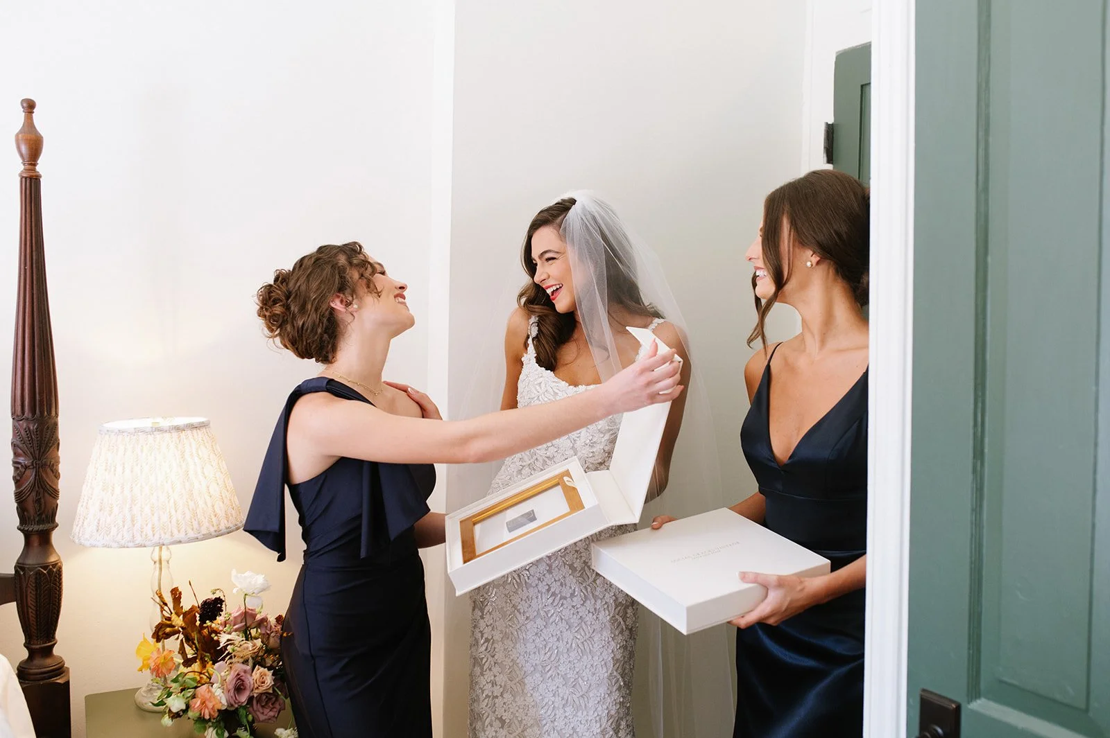 A bride in a white lace wedding dress and veil smiling as she is about to open a gift box from two women in navy blue dresses, one of whom is holding a white box, inside a cozy room with a lamp and flower arrangement.