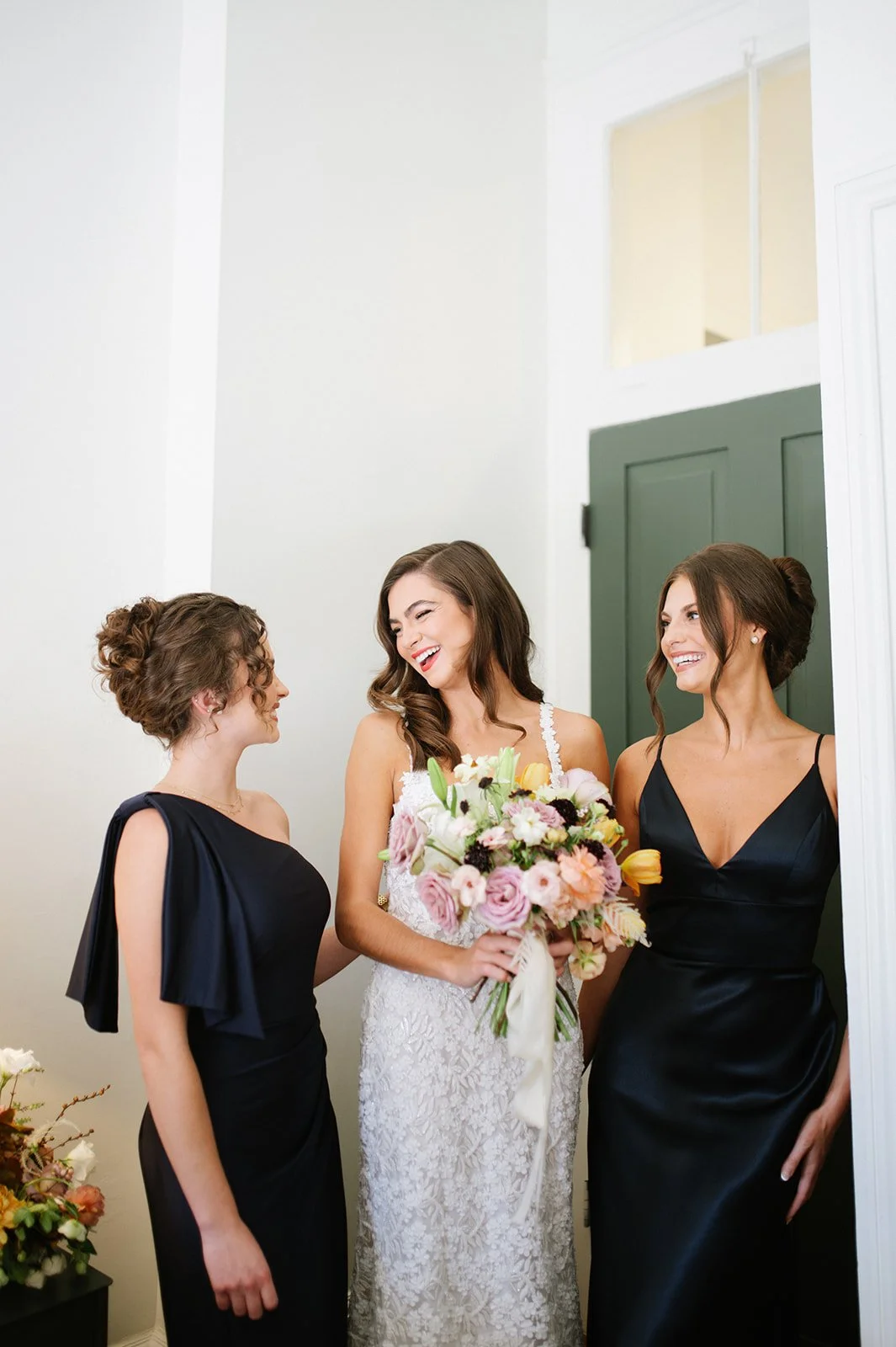 Three women, including a bride with a bouquet, are smiling and talking in a room before a wedding ceremony.