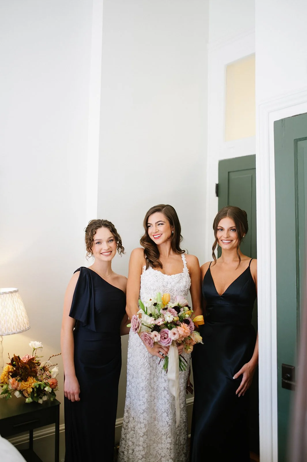 A bride with dark brown hair in a white lace wedding dress holding a bouquet of pink, yellow, and white flowers, standing with two bridesmaids in black dresses with spaghetti straps, all smiling indoors near a green door and a floral arrangement on a