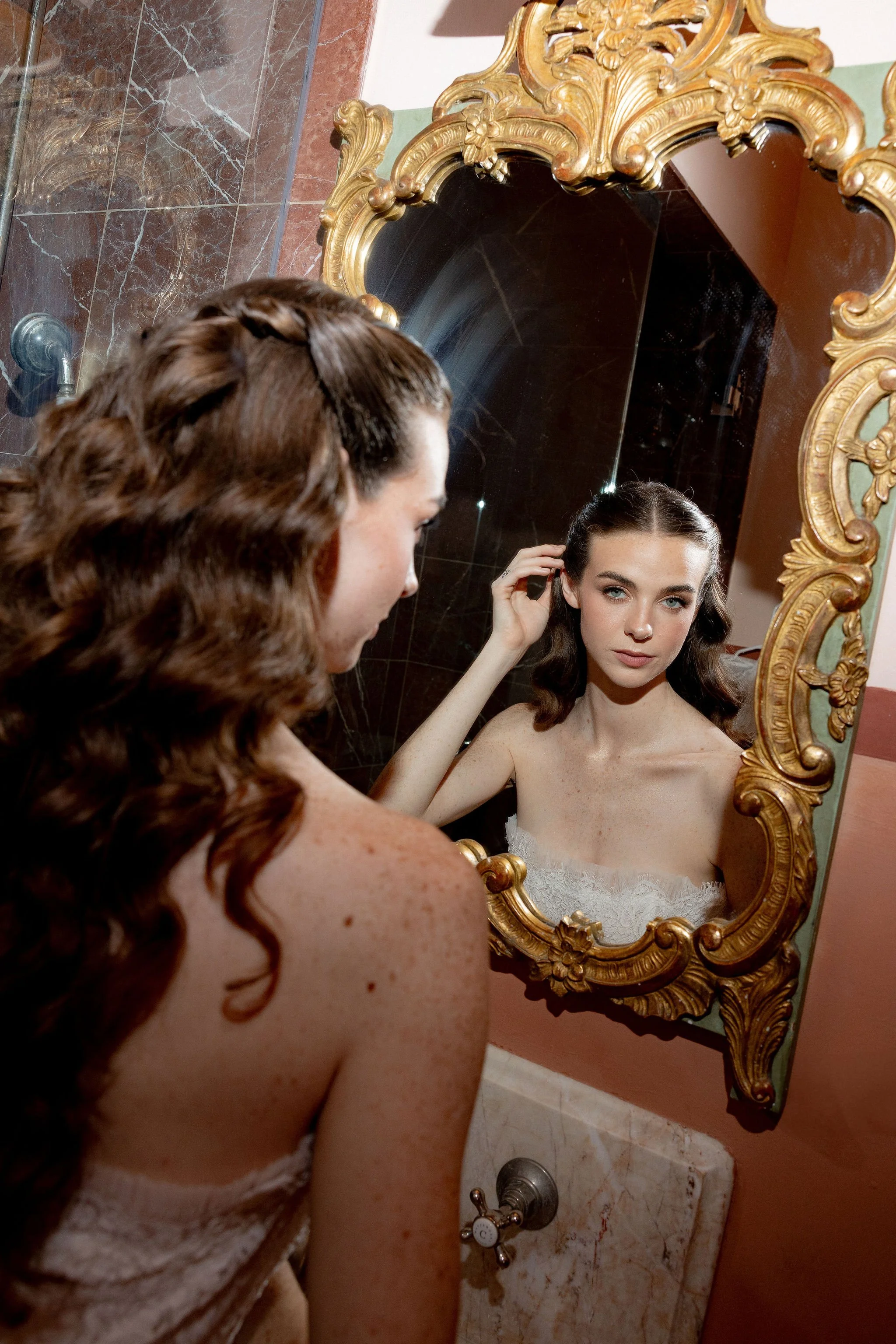 A young woman with dark, wavy hair looking into an ornate gold-framed mirror, touching her hair, with a serious expression on her face. She is in a room with brown and green walls and marble surfaces.